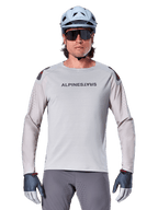 Alpinestars A-Aria Polartec Switch Long Sleeve Jersey, jasnoszara koszulka rowerowa MTB z laserowymi perforacjami na rękawach i logo na klatce piersiowej, noszona z jasnoniebieskim kaskiem, okularami przeciwsłonecznymi i szarymi rękawiczkami