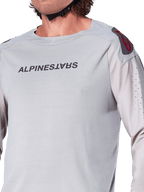 Zbliżenie na jasnoszarą koszulkę z długim rękawem Alpinestars A-Aria Polartec Switch, logo Alpinestars na piersi, czerwone logo na ramieniu i laserowo perforowane rękawy zapewniające przepływ powietrza, wysokiej jakości odzież MTB