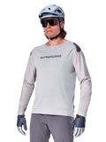 Alpinestars A-Aria Polartec Switch Jersey Long Sleeve, jasnoszara koszulka rowerowa z długim rękawem, czarne logo na klatce piersiowej i czerwony akcent na ramieniu, laserowo perforowane rękawy dla lepszej wentylacji, noszona z jasnoniebieskim kaskiem, okularami przeciwsłonecznymi i szarymi spodniami, profesjonalna odzież MTB na szlaki