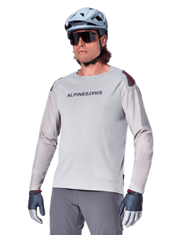 Alpinestars A-Aria Polartec Switch Jersey Long Sleeve, jasnoszara koszulka rowerowa z długim rękawem, czarne logo na klatce piersiowej i czerwony akcent na ramieniu, laserowo perforowane rękawy dla lepszej wentylacji, noszona z jasnoniebieskim kaskiem, okularami przeciwsłonecznymi i szarymi spodniami, profesjonalna odzież MTB na szlaki