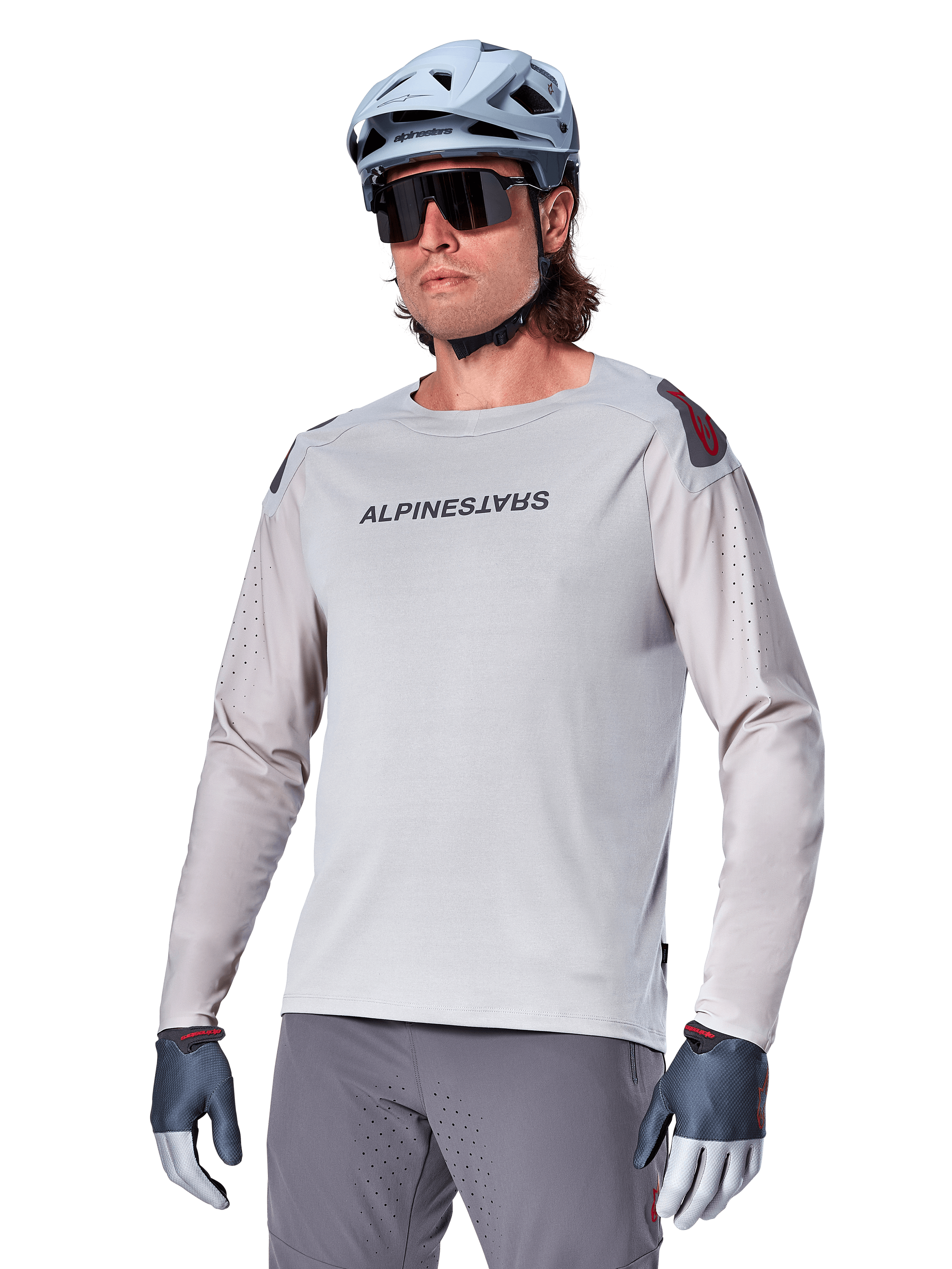 Alpinestars A-Aria Polartec Switch Jersey Long Sleeve, jasnoszara koszulka rowerowa z długim rękawem, czarne logo na klatce piersiowej i czerwony akcent na ramieniu, laserowo perforowane rękawy dla lepszej wentylacji, noszona z jasnoniebieskim kaskiem, okularami przeciwsłonecznymi i szarymi spodniami, profesjonalna odzież MTB na szlaki
