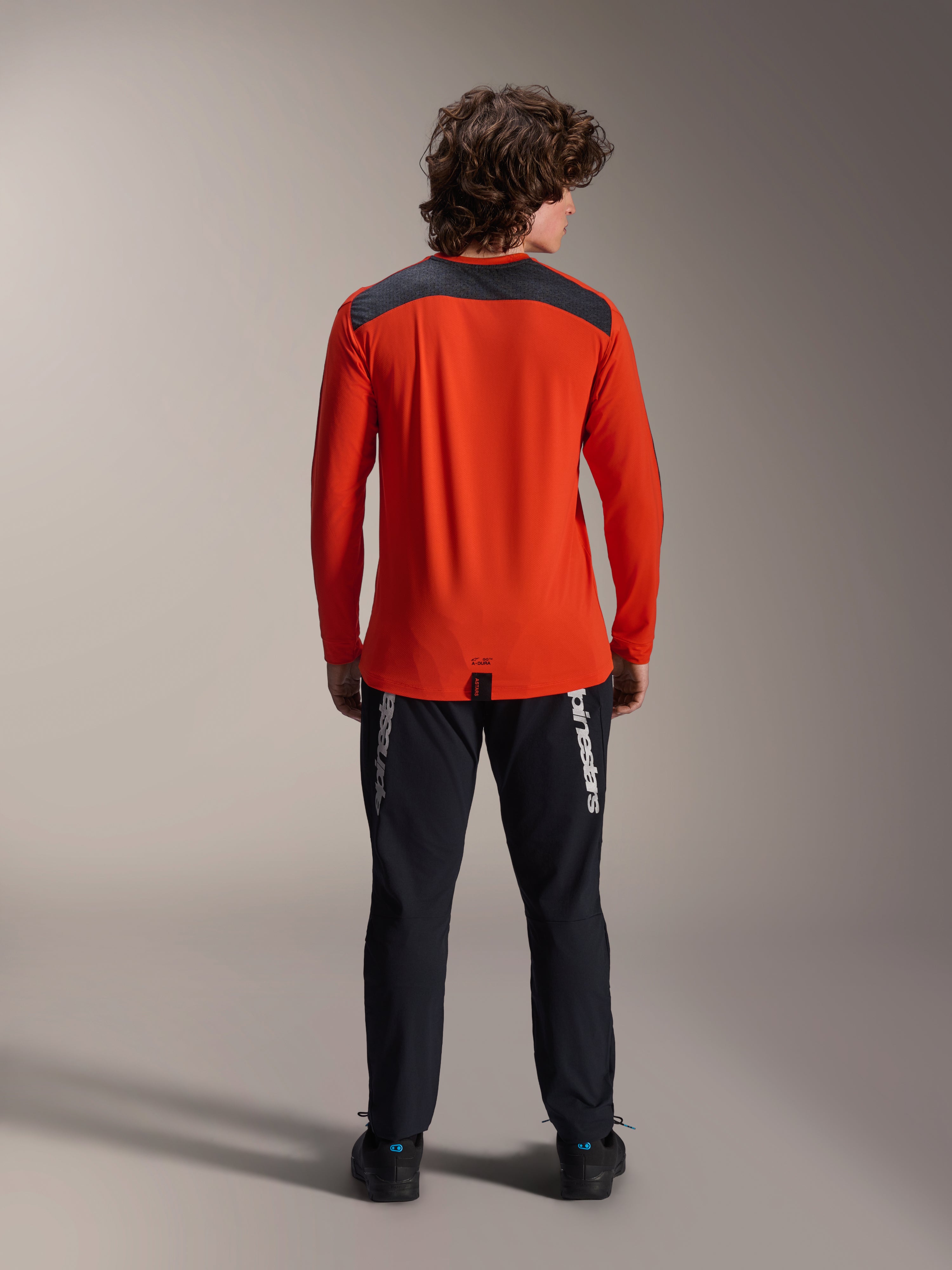 Koszulka z długim rękawem Alpinestars A-Dura Insert Astar w kolorze Spicy Orange, widok z tyłu prezentujący grafitowy panel na ramionach, noszona z czarnymi spodniami Alpinestars i czarnymi butami, profesjonalne zdjęcie studyjne odzieży MTB