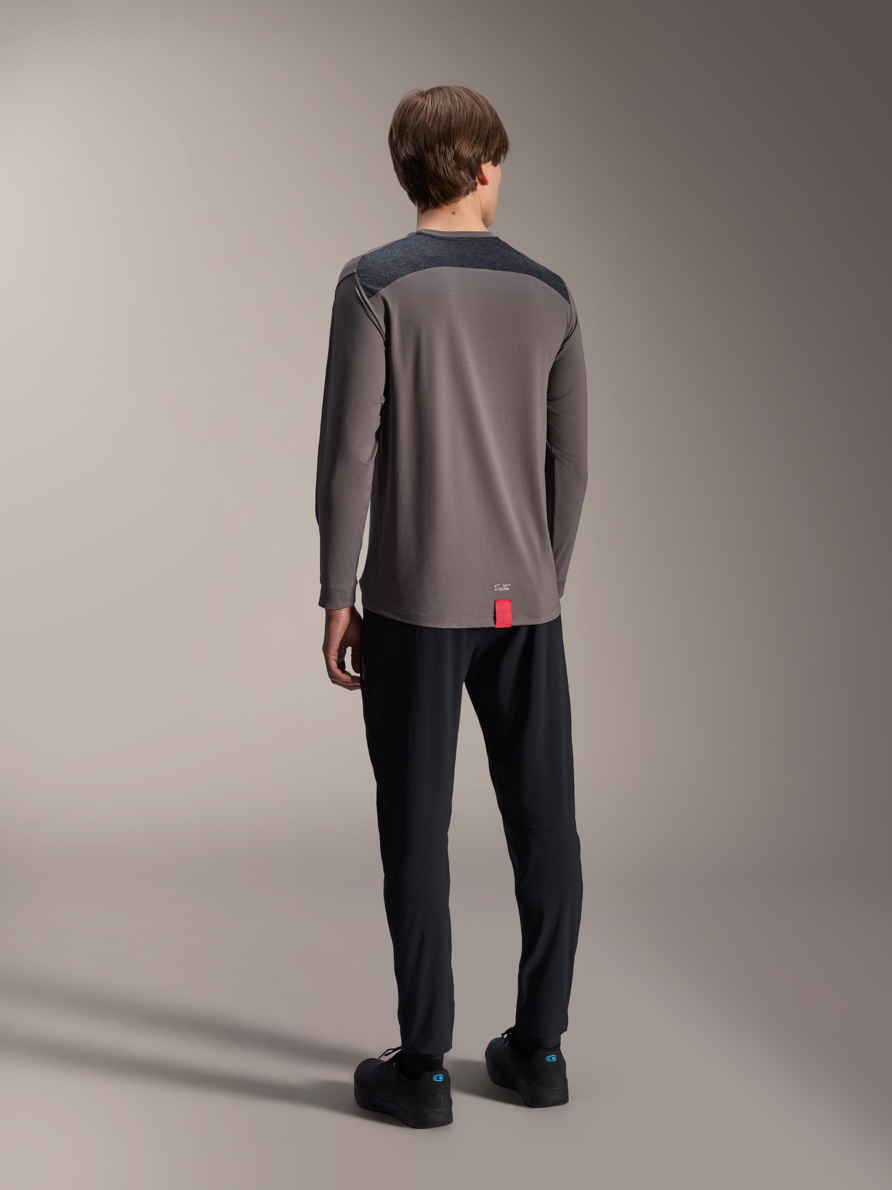 Alpinestars A-Dura Insert Astar Long Sleeve Jersey, ciemnoszara koszulka z długim rękawem, widok z tyłu na modelu, grafitowe wzmocnione panele na ramionach i mała czerwona metka u dołu, w zestawieniu z czarnymi spodniami i butami, techniczna odzież MTB
