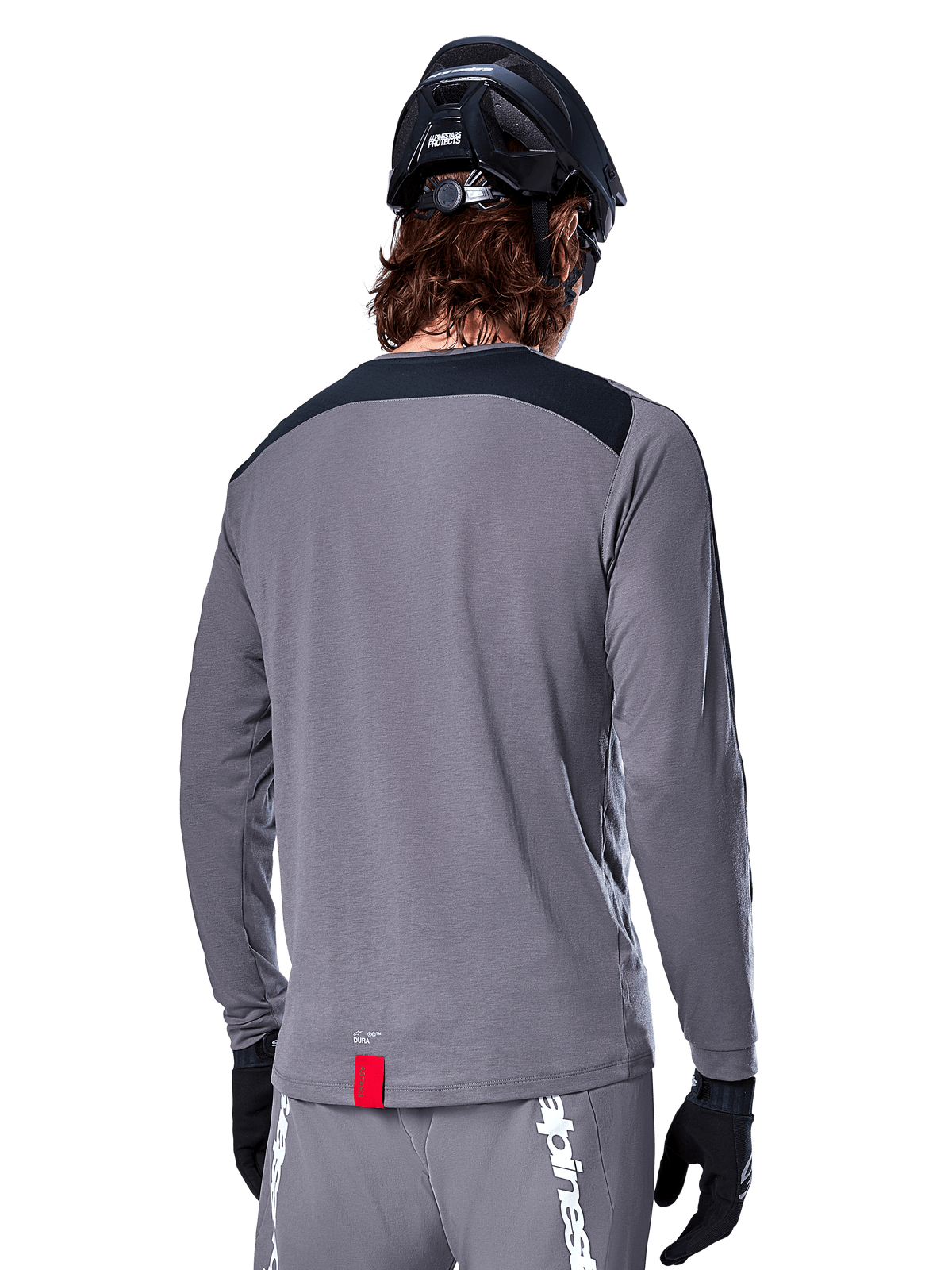 Koszulka Alpinestars A-Dura Dri Astar Long Sleeve, ciemnoszara koszulka MTB widoczna od tyłu, czarny panel na ramionach, mała czerwona metka, czarny kask i rękawiczki rowerowe