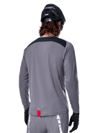Koszulka Alpinestars A-Dura Dri Astar Long Sleeve, ciemnoszara koszulka MTB widoczna od tyłu, czarny panel na ramionach, mała czerwona metka, czarny kask i rękawiczki rowerowe