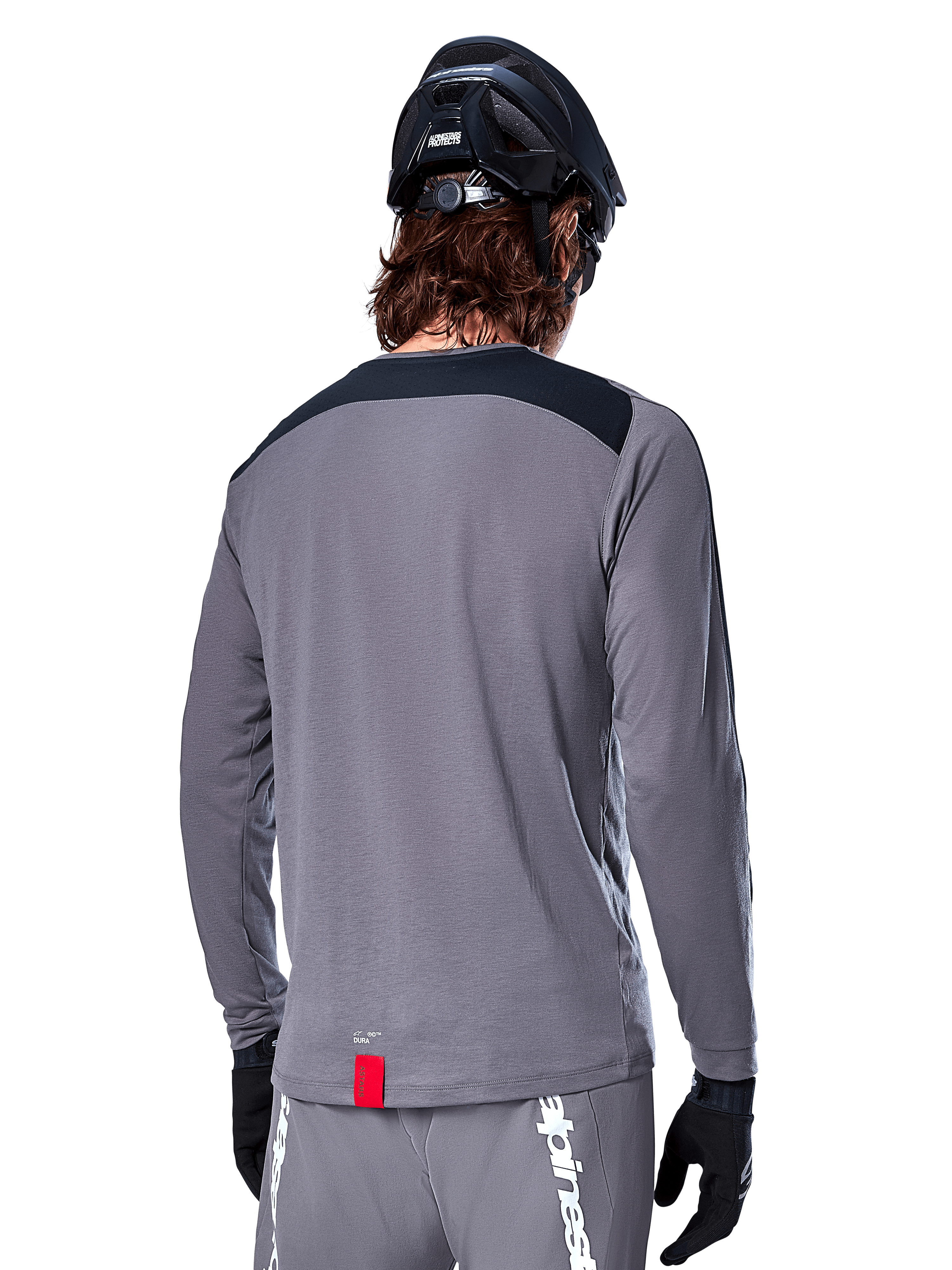Koszulka Alpinestars A-Dura Dri Astar Long Sleeve, ciemnoszara koszulka MTB widoczna od tyłu, czarny panel na ramionach, mała czerwona metka, czarny kask i rękawiczki rowerowe