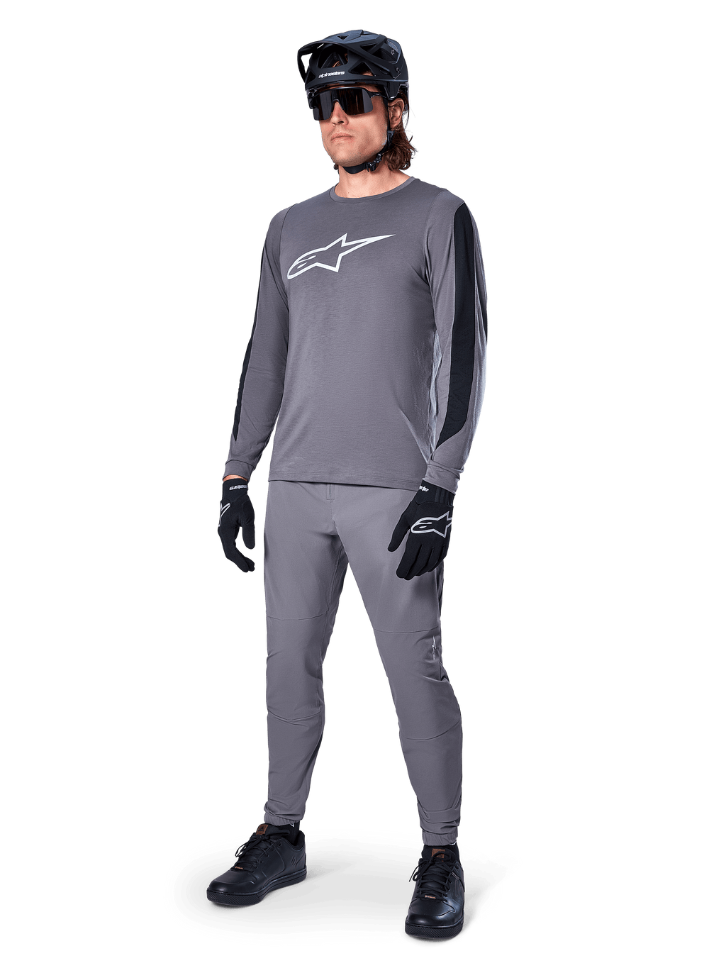 Koszulka Alpinestars A-Dura Dri Astar, koszulka MTB z długim rękawem, ciemnoszara z białym logo na piersi, czarne panele z siatki na rękawach, zestawiona z pasującymi szarymi spodniami, czarnym kaskiem i rękawiczkami, wyczynowa odzież do kolarstwa górskiego