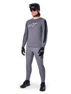 Koszulka Alpinestars A-Dura Dri Astar, koszulka MTB z długim rękawem, ciemnoszara z białym logo na piersi, czarne panele z siatki na rękawach, zestawiona z pasującymi szarymi spodniami, czarnym kaskiem i rękawiczkami, wyczynowa odzież do kolarstwa górskiego