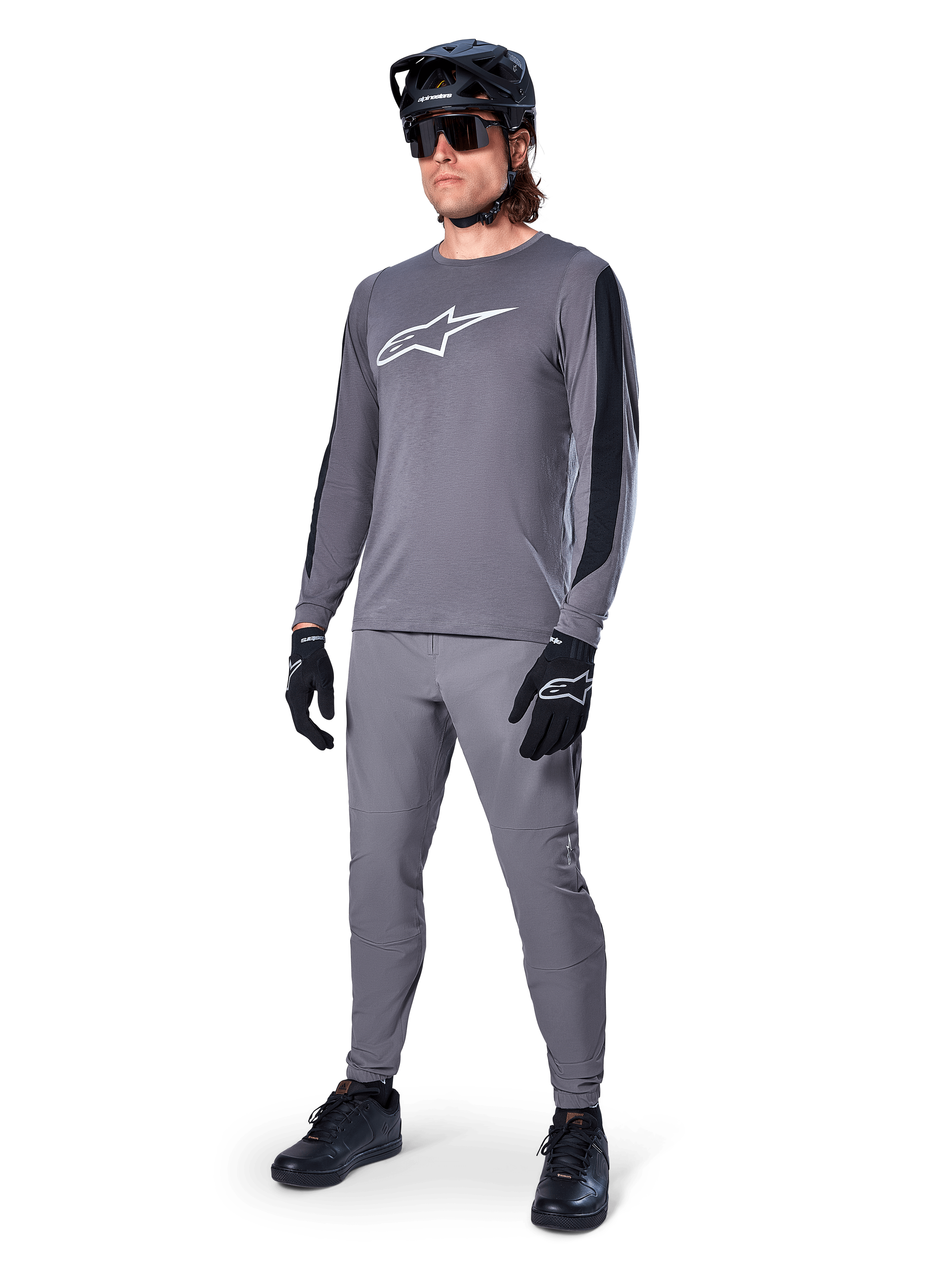 Koszulka Alpinestars A-Dura Dri Astar, koszulka MTB z długim rękawem, ciemnoszara z białym logo na piersi, czarne panele z siatki na rękawach, zestawiona z pasującymi szarymi spodniami, czarnym kaskiem i rękawiczkami, wyczynowa odzież do kolarstwa górskiego