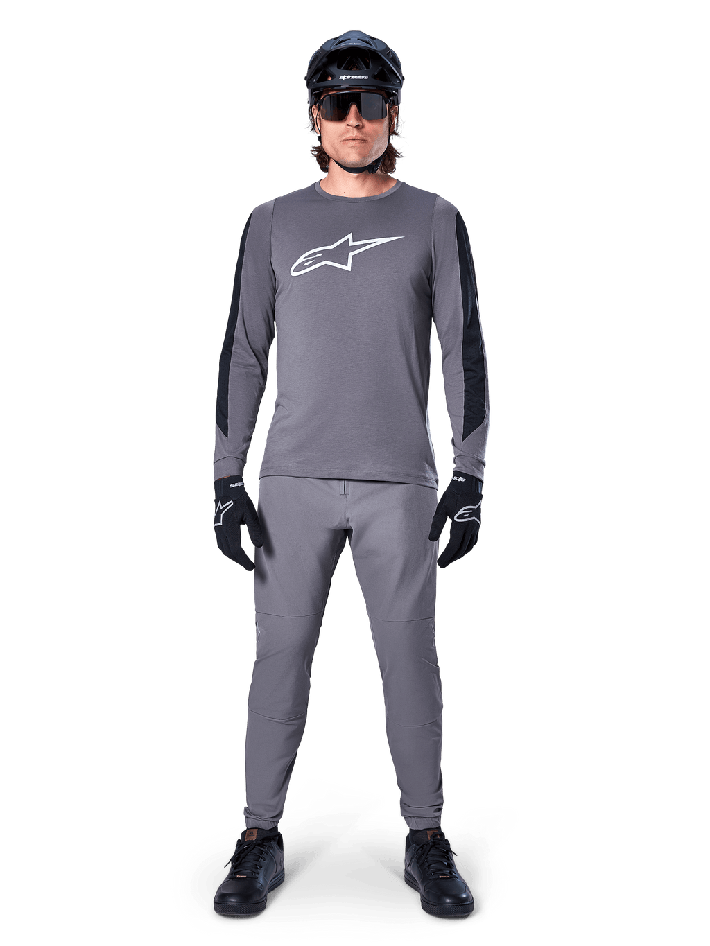 Alpinestars A-Dura Dri Astar Jersey, koszulka MTB z długim rękawem w kolorze ciemnoszarym, z białym logo na piersi i czarnymi panelami na rękawach, noszona z pasującymi szarymi spodniami, czarnym kaskiem, okularami przeciwsłonecznymi i rękawiczkami, tworząca kompletny strój do jazdy terenowej