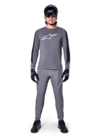 Alpinestars A-Dura Dri Astar Jersey, koszulka MTB z długim rękawem w kolorze ciemnoszarym, z białym logo na piersi i czarnymi panelami na rękawach, noszona z pasującymi szarymi spodniami, czarnym kaskiem, okularami przeciwsłonecznymi i rękawiczkami, tworząca kompletny strój do jazdy terenowej