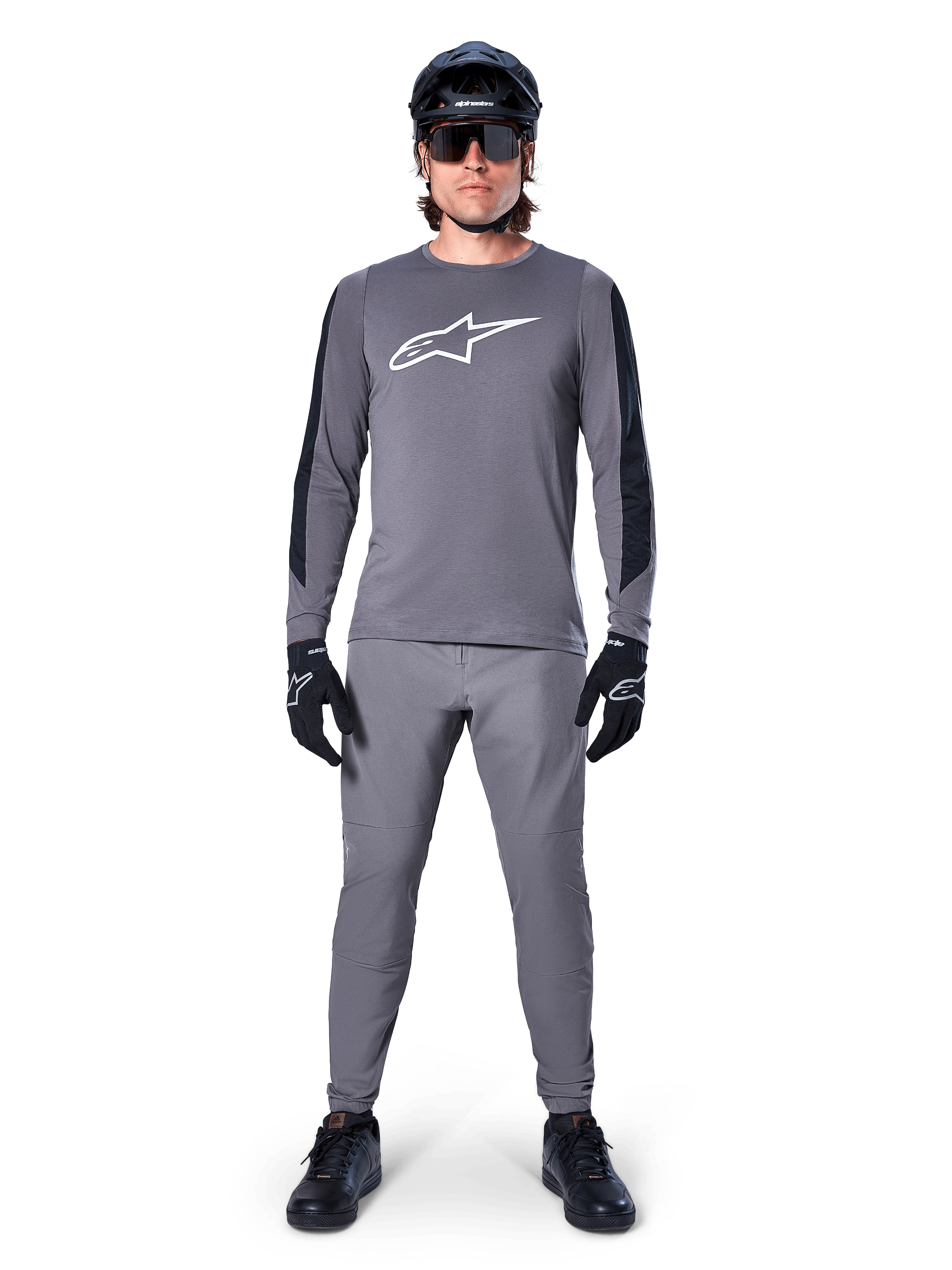 Alpinestars A-Dura Dri Astar Jersey, koszulka MTB z długim rękawem w kolorze ciemnoszarym, z białym logo na piersi i czarnymi panelami na rękawach, noszona z pasującymi szarymi spodniami, czarnym kaskiem, okularami przeciwsłonecznymi i rękawiczkami, tworząca kompletny strój do jazdy terenowej