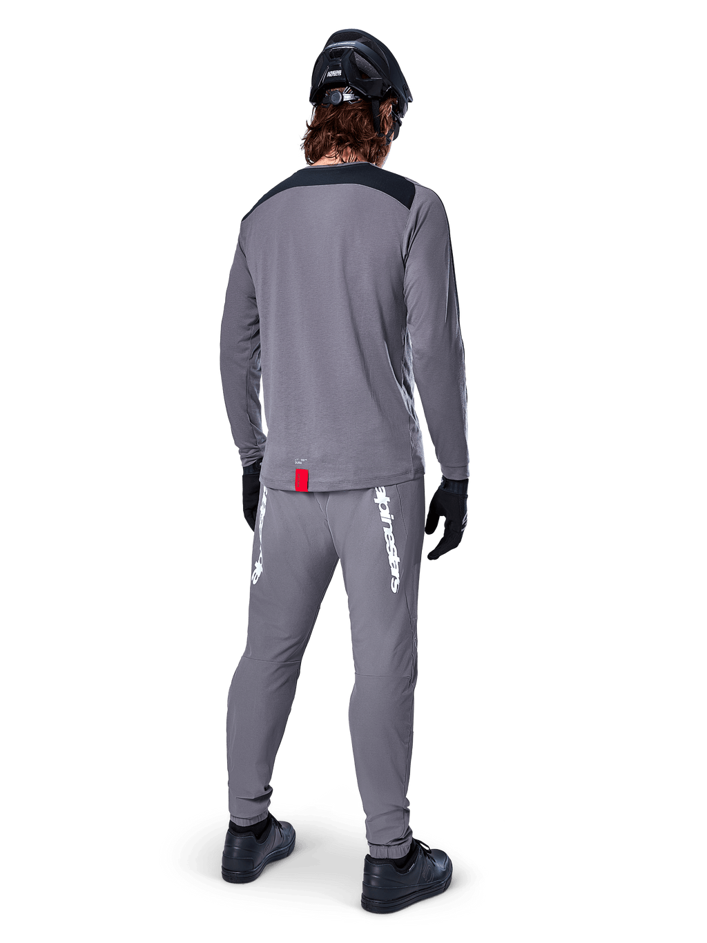 Alpinestars A-Dura Dri Astar Jersey - Long Sleeve, ciemnoszara, widok od tyłu na rowerzystę w pasujących spodniach i czarnym kasku, posiada czarne panele na ramionach i małą czerwoną metkę u dołu, techniczna odzież do jazdy terenowej