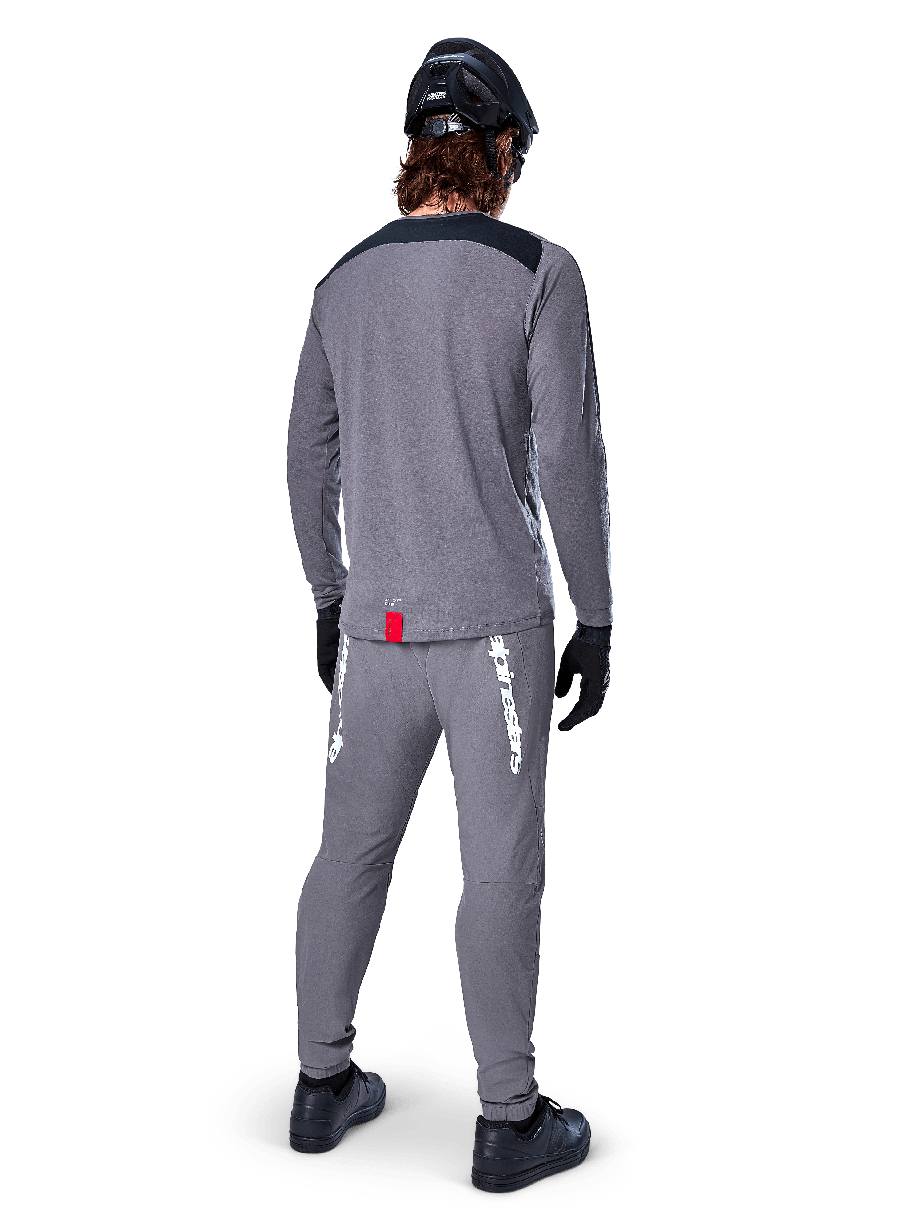 Alpinestars A-Dura Dri Astar Jersey - Long Sleeve, ciemnoszara, widok od tyłu na rowerzystę w pasujących spodniach i czarnym kasku, posiada czarne panele na ramionach i małą czerwoną metkę u dołu, techniczna odzież do jazdy terenowej