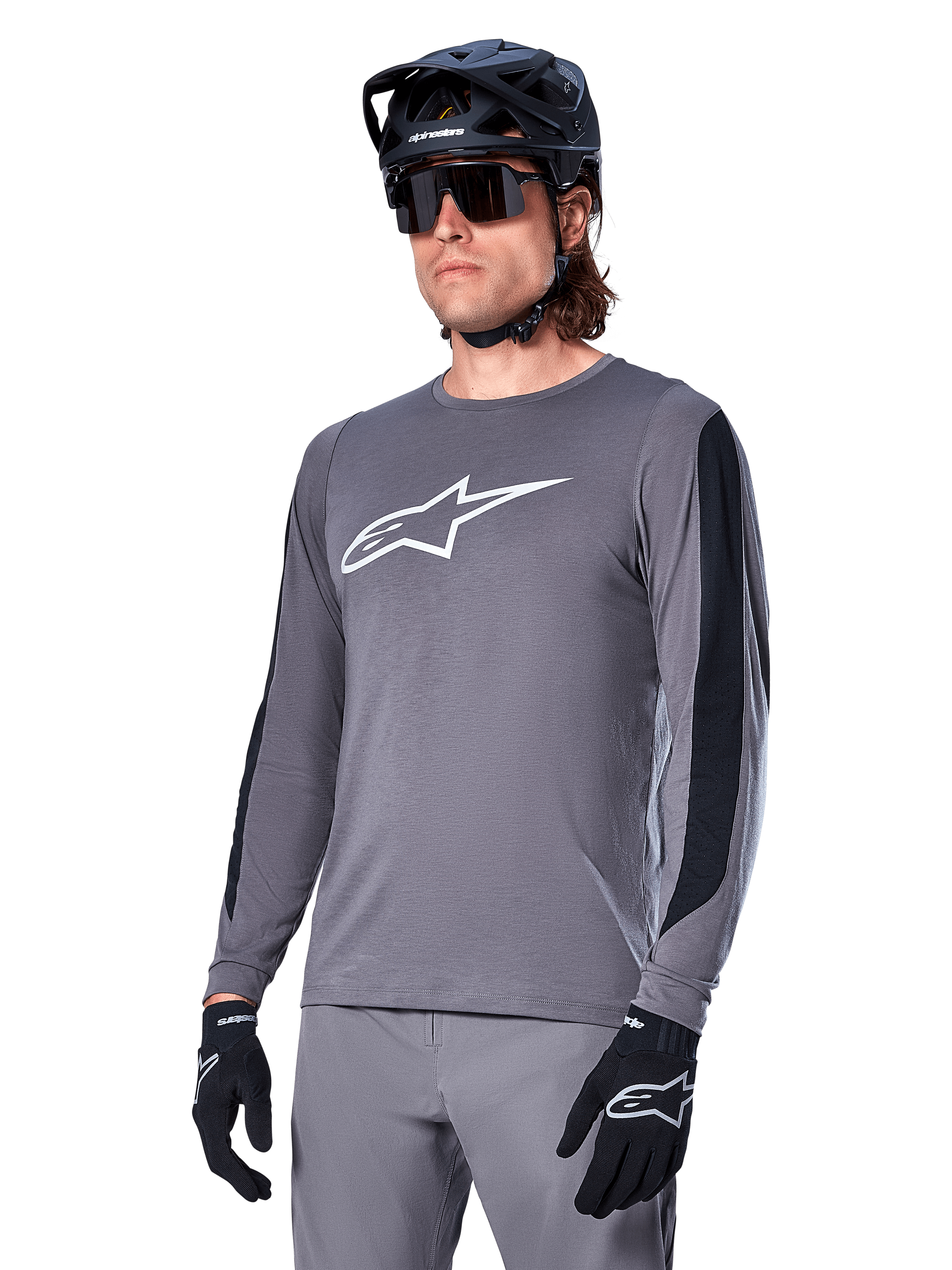 Alpinestars A-Dura Dri Astar Jersey Long Sleeve, ciemnoszara koszulka rowerowa MTB, białe logo na klatce piersiowej i czarne siatkowe panele boczne, noszona z czarnym kaskiem, okularami przeciwsłonecznymi i pasującymi szarymi spodniami