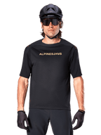 Rowerzysta górski w koszulce Alpinestars A-Aria Polartec Switch z krótkim rękawem w kolorze czarnym, ze złotym brandingiem na klatce piersiowej, w zestawie z czarnym kaskiem MTB, ciemnymi okularami sportowymi i pasującymi rękawiczkami, odzież do jazdy terenowej.