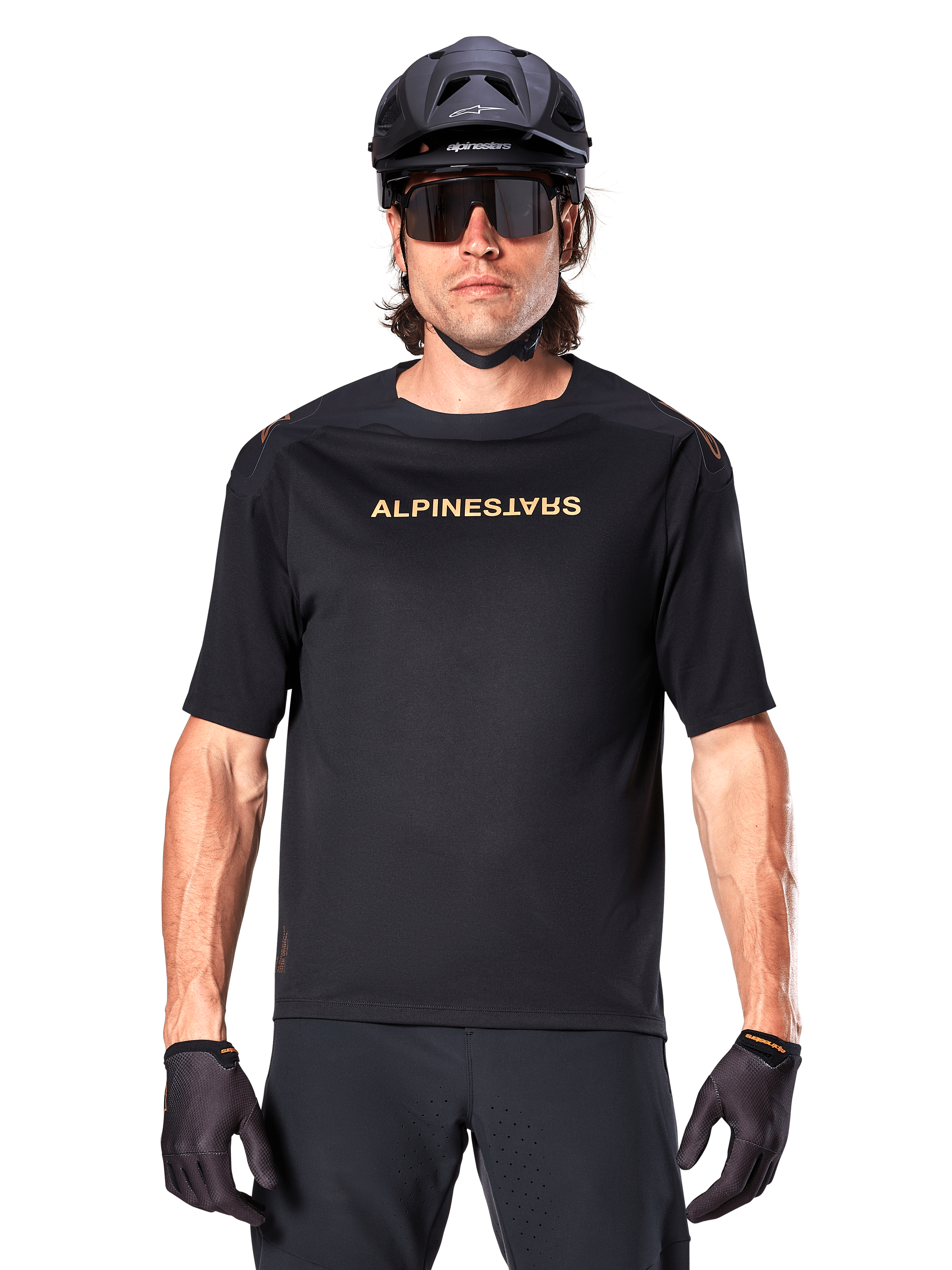Rowerzysta górski w koszulce Alpinestars A-Aria Polartec Switch z krótkim rękawem w kolorze czarnym, ze złotym brandingiem na klatce piersiowej, w zestawie z czarnym kaskiem MTB, ciemnymi okularami sportowymi i pasującymi rękawiczkami, odzież do jazdy terenowej.