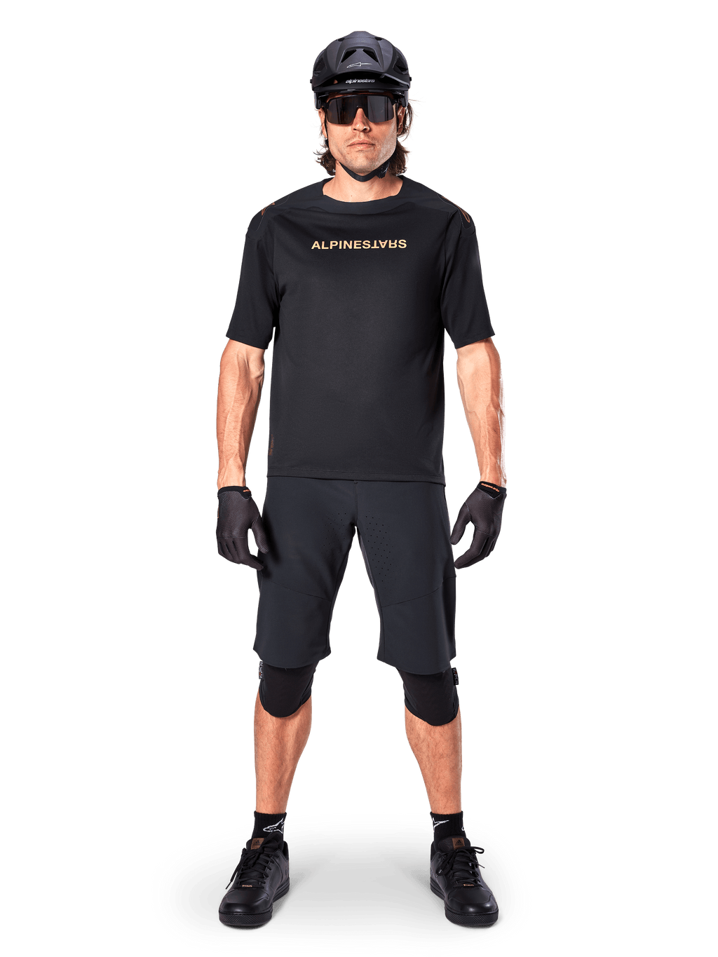 Alpinestars A-Aria Polartec Switch Jersey, koszulka MTB z krótkim rękawem, czarna z beżowymi napisami, noszona z dopasowanym czarnym ekwipunkiem do kolarstwa górskiego, w tym kaskiem, szortami i ochraniaczami na kolana, techniczna odzież rowerowa do jazdy w terenie