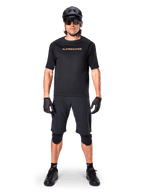 Alpinestars A-Aria Polartec Switch Jersey, koszulka MTB z krótkim rękawem, czarna z beżowymi napisami, noszona z dopasowanym czarnym ekwipunkiem do kolarstwa górskiego, w tym kaskiem, szortami i ochraniaczami na kolana, techniczna odzież rowerowa do jazdy w terenie