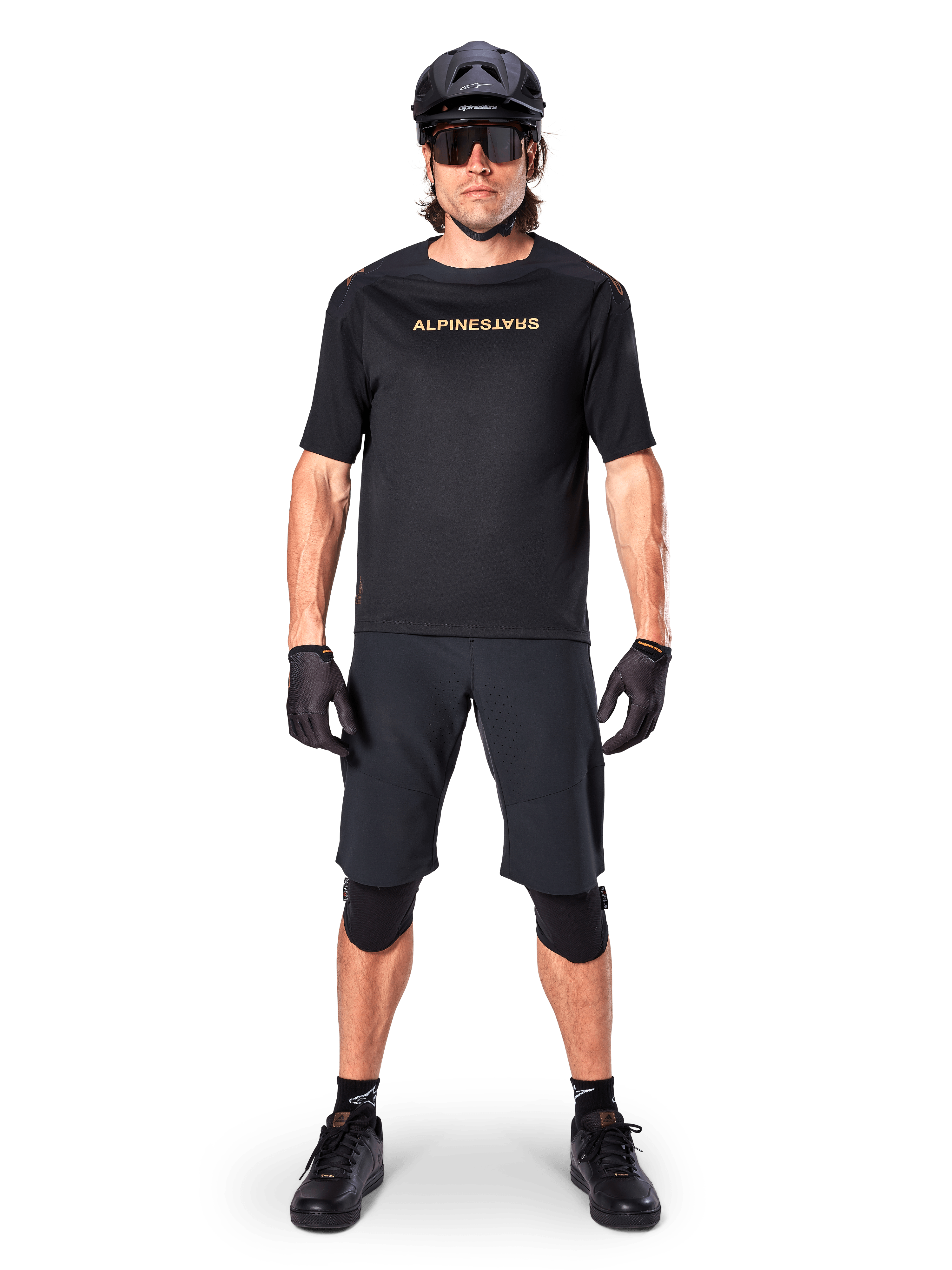 Alpinestars A-Aria Polartec Switch Jersey, koszulka MTB z krótkim rękawem, czarna z beżowymi napisami, noszona z dopasowanym czarnym ekwipunkiem do kolarstwa górskiego, w tym kaskiem, szortami i ochraniaczami na kolana, techniczna odzież rowerowa do jazdy w terenie