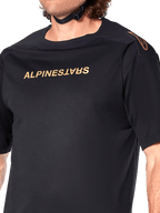 Alpinestars A-Aria Polartec Switch Jersey, koszulka MTB z krótkim rękawem, czarna, złoty napis Alpinestars na klatce piersiowej i logo na ramieniu, zbliżenie na górną część tułowia podkreślające techniczny materiał i sportowy krój