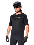 Alpinestars A-Aria Polartec Switch Jersey z krótkim rękawem, czarna koszulka rowerowa ze złotym brandingiem Alpinestars, czarny kask, okulary przeciwsłoneczne i rękawice, techniczna odzież MTB