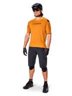 Male rider wearing the Alpinestars A-Aria Polartec Switch Jersey in Dark Gold, short-sleeve MTB jersey, paired with black cycling shorts, knee guards, black helmet, and gloves, professional mountain bike outfit for trail riding