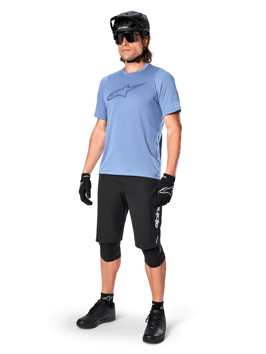 Rowerzysta w koszulce z krótkim rękawem Alpinestars A-Dura Dri Astar w kolorze Infinity Blue, czarne spodenki MTB, czarny kask, okulary przeciwsłoneczne, ochraniacze na kolana i rękawiczki, kompletny strój do kolarstwa górskiego