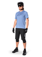 Rowerzysta w koszulce z krótkim rękawem Alpinestars A-Dura Dri Astar w kolorze Infinity Blue, czarne spodenki MTB, czarny kask, okulary przeciwsłoneczne, ochraniacze na kolana i rękawiczki, kompletny strój do kolarstwa górskiego