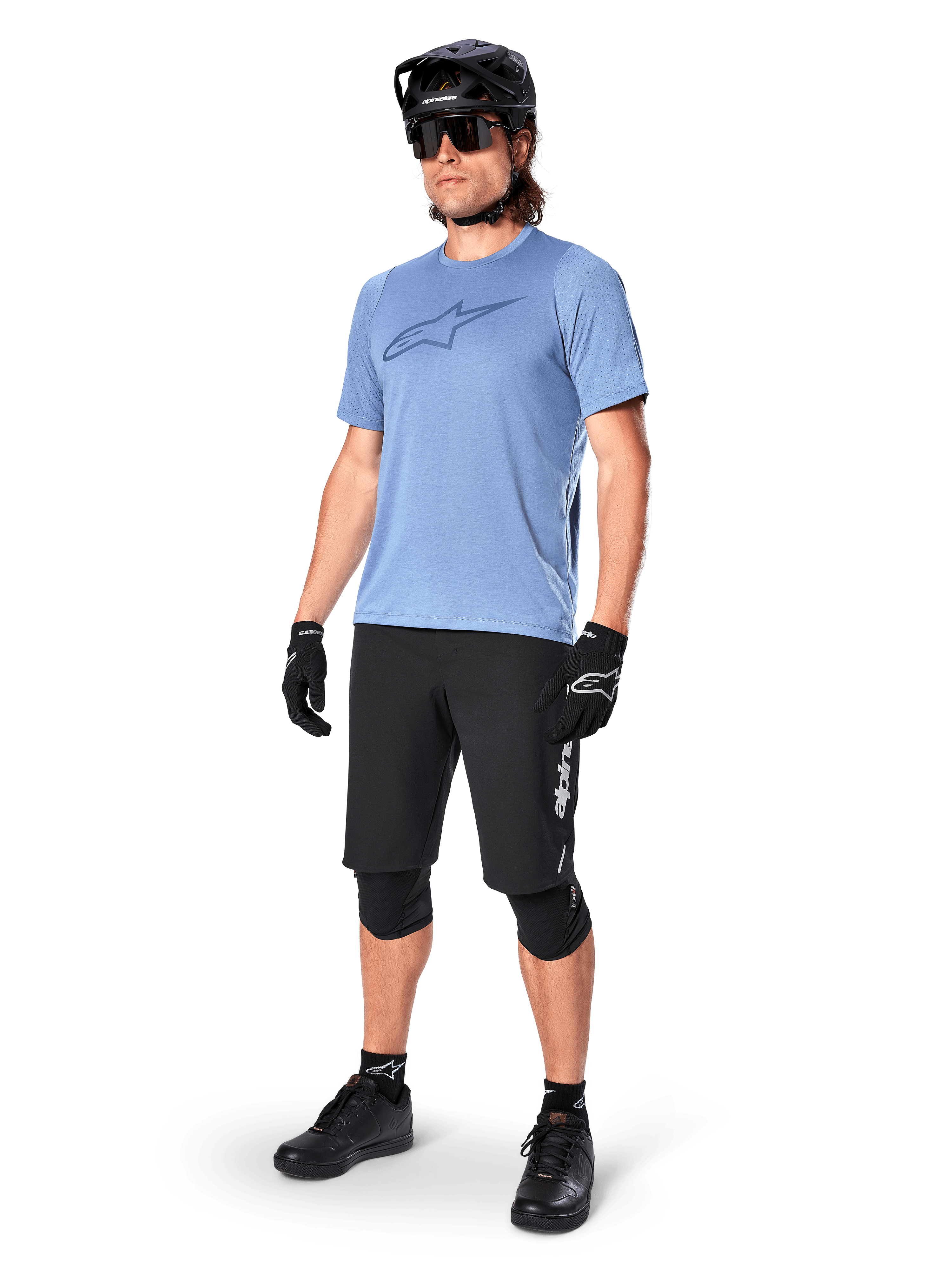 Rowerzysta w koszulce z krótkim rękawem Alpinestars A-Dura Dri Astar w kolorze Infinity Blue, czarne spodenki MTB, czarny kask, okulary przeciwsłoneczne, ochraniacze na kolana i rękawiczki, kompletny strój do kolarstwa górskiego