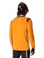 Alpinestars A-Dura Dri Wool Long Sleeve Jersey w kolorze Dark Gold, widok z tyłu na rowerzystę w kasku MTB i rękawiczkach, czarne panele z siatki na ramionach i przedłużony tył, techniczna odzież rowerowa do jazdy po szlakach