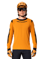 Męski model w koszulce z długim rękawem Alpinestars A-Dura Dri Wool w kolorze Dark Gold, dopasowany złoty kask MTB i rękawice, czarne panele z siatki na ramionach i rękawach, czarny napis Alpinestars na piersi, ciemne okulary przeciwsłoneczne i spodnie, techniczna odzież do kolarstwa górskiego