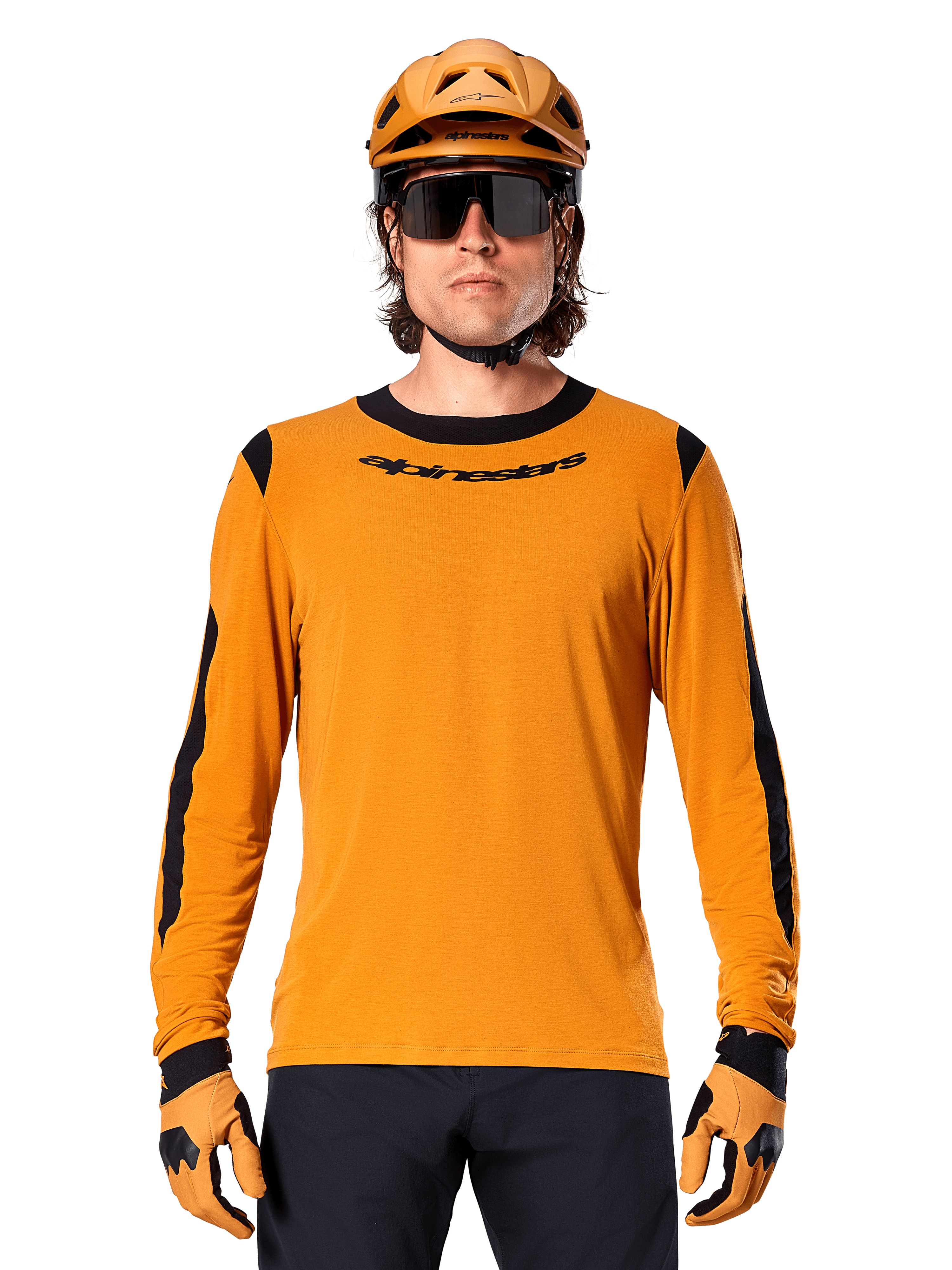 Męski model w koszulce z długim rękawem Alpinestars A-Dura Dri Wool w kolorze Dark Gold, dopasowany złoty kask MTB i rękawice, czarne panele z siatki na ramionach i rękawach, czarny napis Alpinestars na piersi, ciemne okulary przeciwsłoneczne i spodnie, techniczna odzież do kolarstwa górskiego