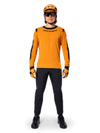 Rowerzysta górski w koszulce Alpinestars A-Dura Dri Wool Long Sleeve Jersey, ciemnozłota z czarnymi akcentami na kołnierzu i rękawach, czarne spodnie, pasujący złoty kask, rękawice i czarne okulary przeciwsłoneczne, profesjonalny sprzęt do kolarstwa górskiego