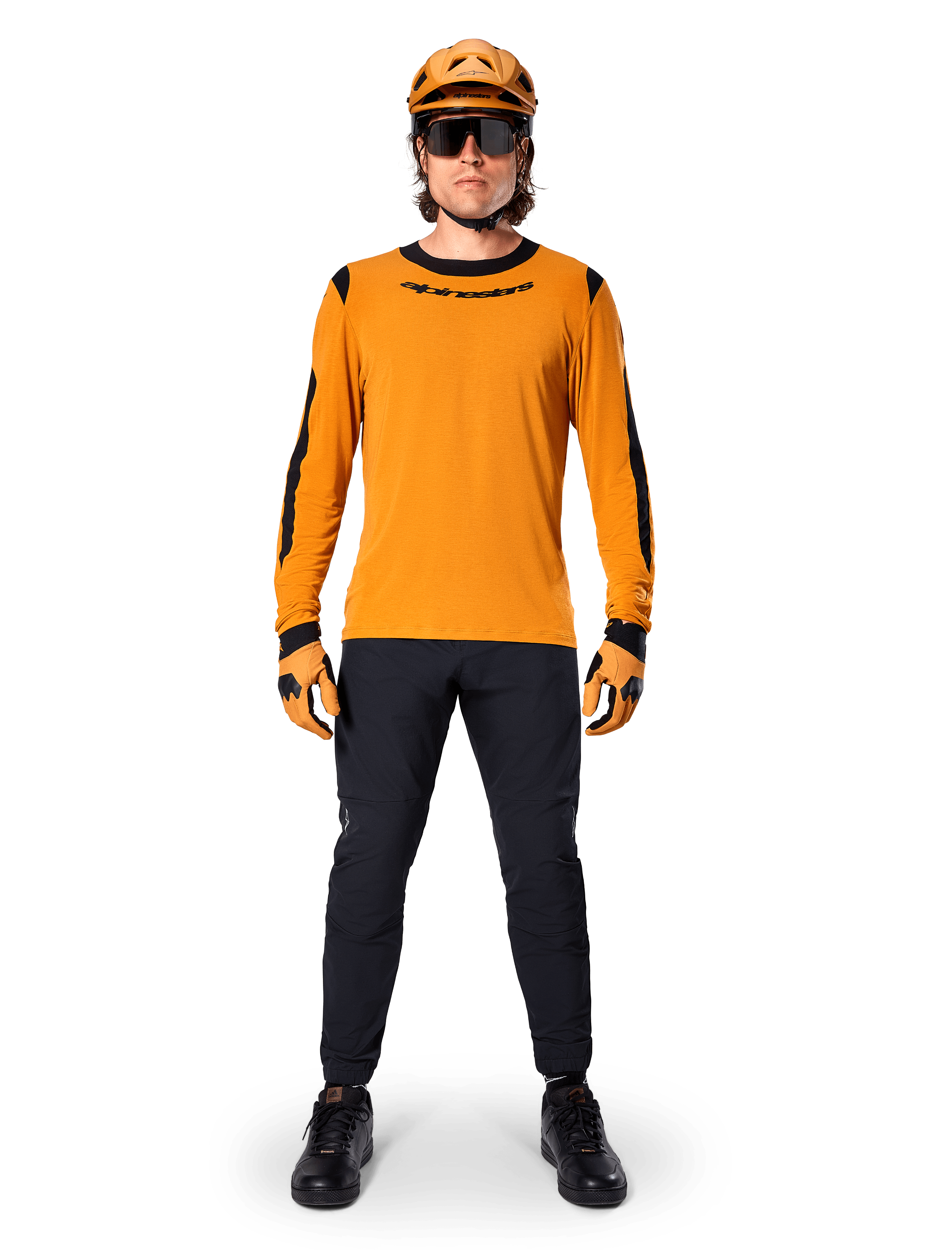 Rowerzysta górski w koszulce Alpinestars A-Dura Dri Wool Long Sleeve Jersey, ciemnozłota z czarnymi akcentami na kołnierzu i rękawach, czarne spodnie, pasujący złoty kask, rękawice i czarne okulary przeciwsłoneczne, profesjonalny sprzęt do kolarstwa górskiego