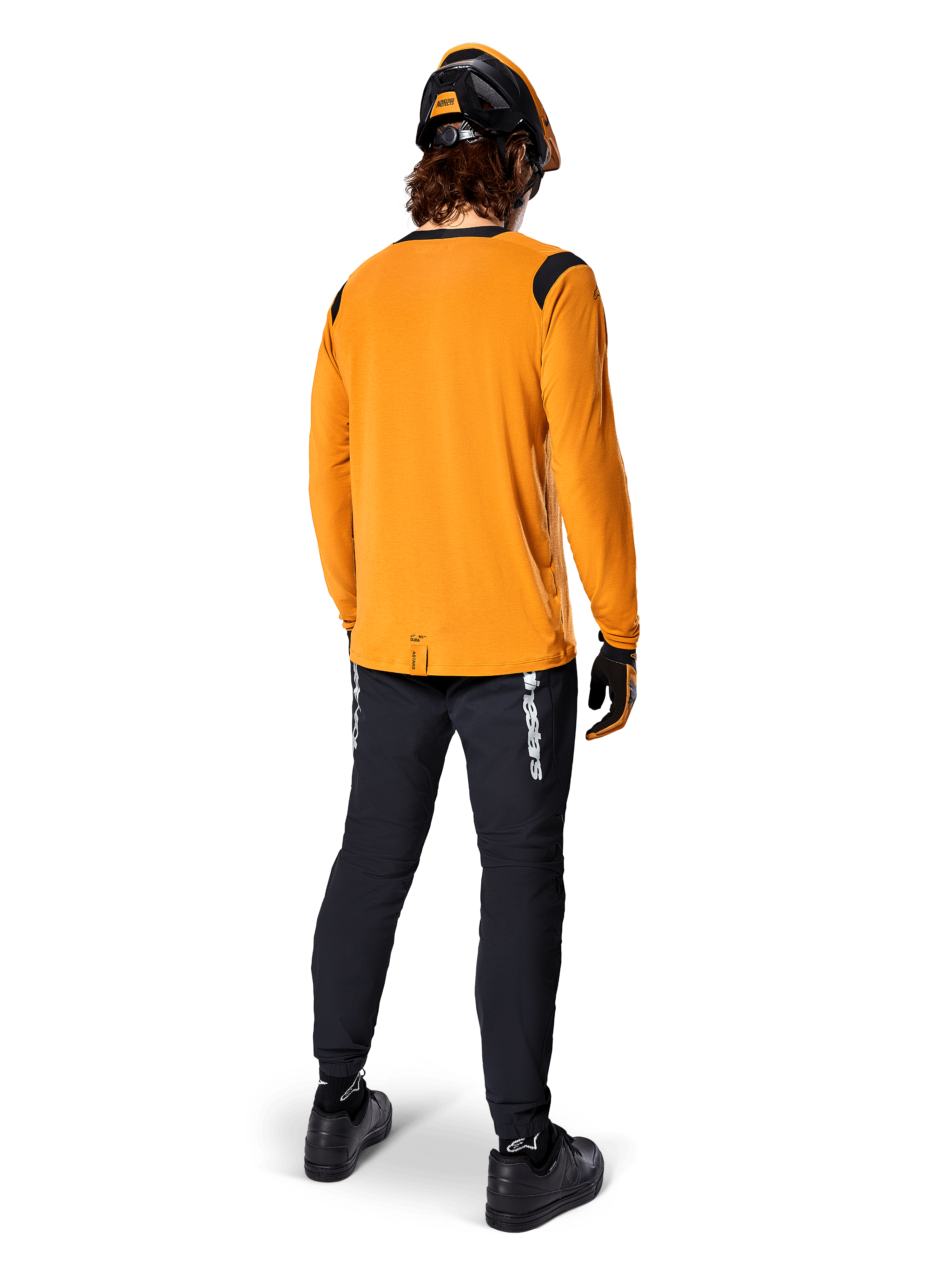 Rowerzysta górski widziany od tyłu w koszulce Alpinestars A-Dura Dri Wool z długim rękawem w kolorze Dark Gold, czarne spodnie techniczne z białym logo i dopasowany czarno-pomarańczowy kask, profesjonalna odzież do kolarstwa górskiego