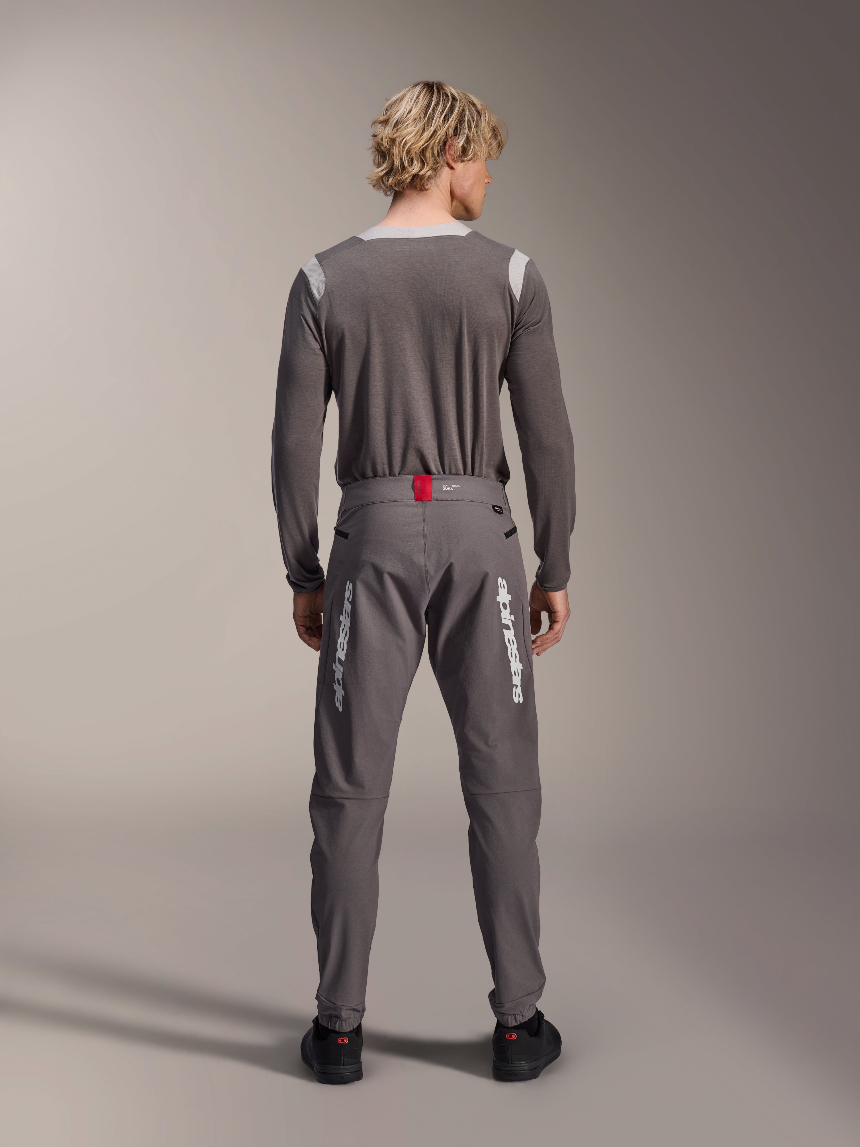 Model widoczny od tyłu w spodniach Alpinestars A-Dura Elite w kolorze ciemnoszarym i pasującej szarej koszulce z długim rękawem, białe logo Alpinestars na nogawkach, czerwony akcent w pasie, czarne buty, profesjonalna odzież rowerowa MTB.