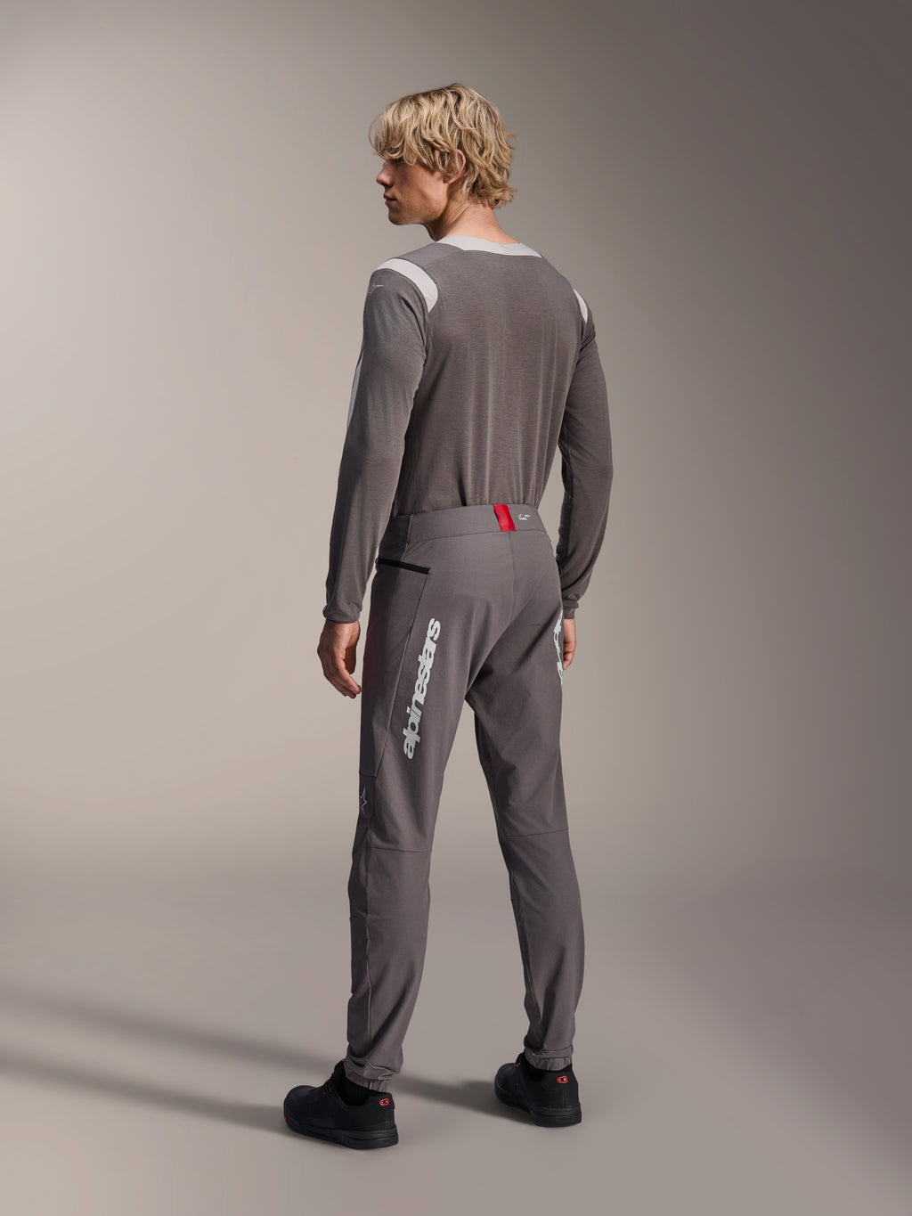 Model w widoku od tyłu ubrany w odzież MTB Alpinestars, szara koszulka z długim rękawem A-Dura Dri Wool, ciemnoszare spodnie A-Dura Elite z białym pionowym brandingiem i czerwonym detalem w pasie, dopasowany strój do kolarstwa górskiego