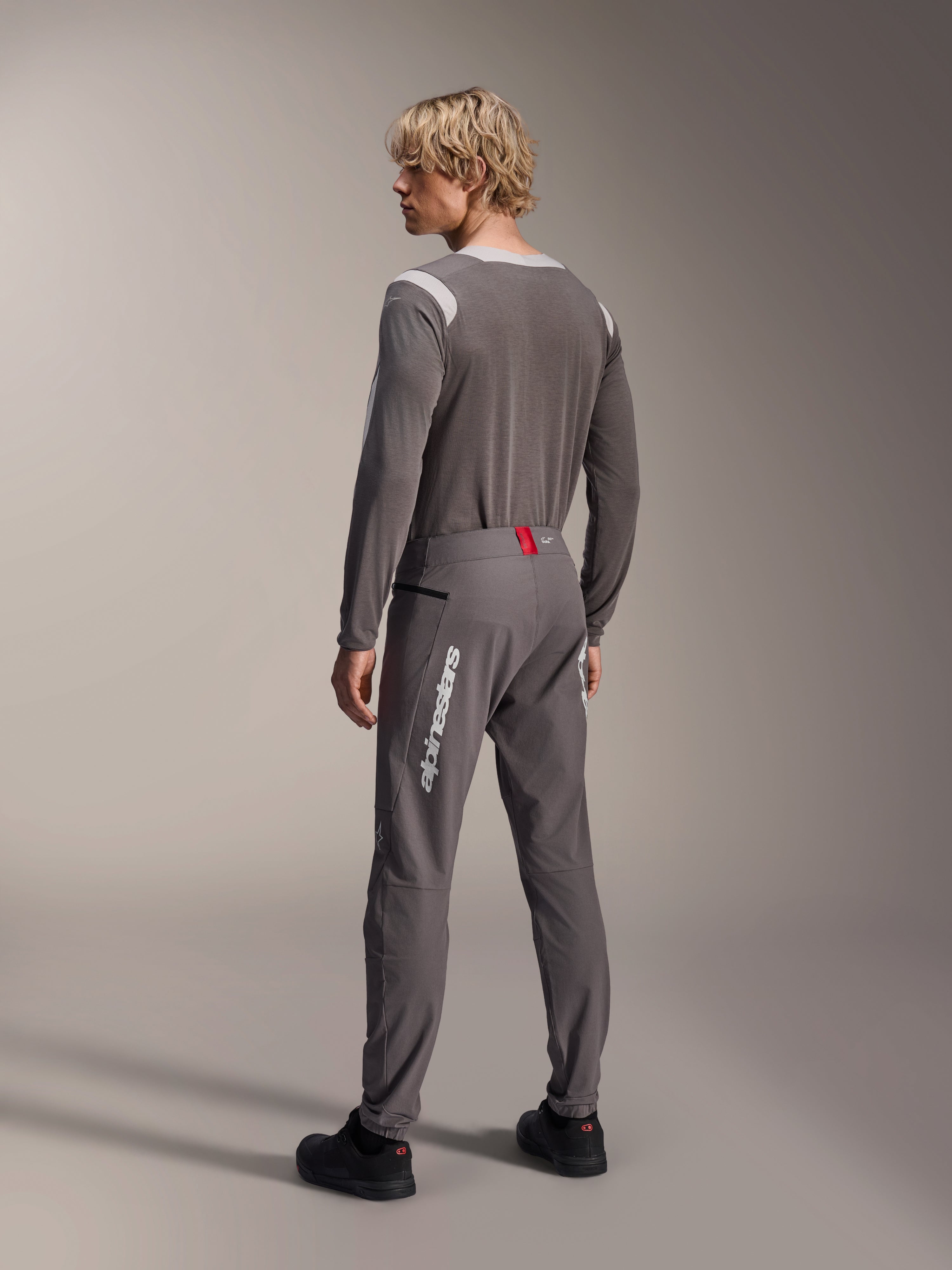 Model w widoku od tyłu ubrany w odzież MTB Alpinestars, szara koszulka z długim rękawem A-Dura Dri Wool, ciemnoszare spodnie A-Dura Elite z białym pionowym brandingiem i czerwonym detalem w pasie, dopasowany strój do kolarstwa górskiego