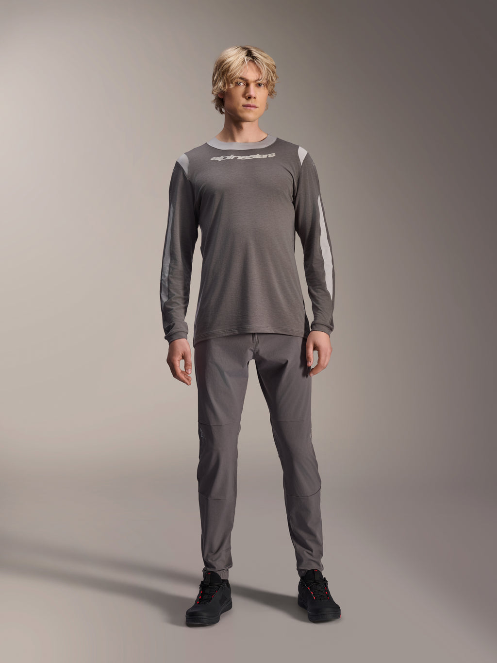 Alpinestars A-Dura Dri Wool Long Sleeve Jersey, ciemnoszara, model w pasujących szarych spodniach i czarnych butach, białe logo Alpinestars na piersi, jasnoszare panele z siatki na ramionach i rękawach, techniczna odzież na rower górski