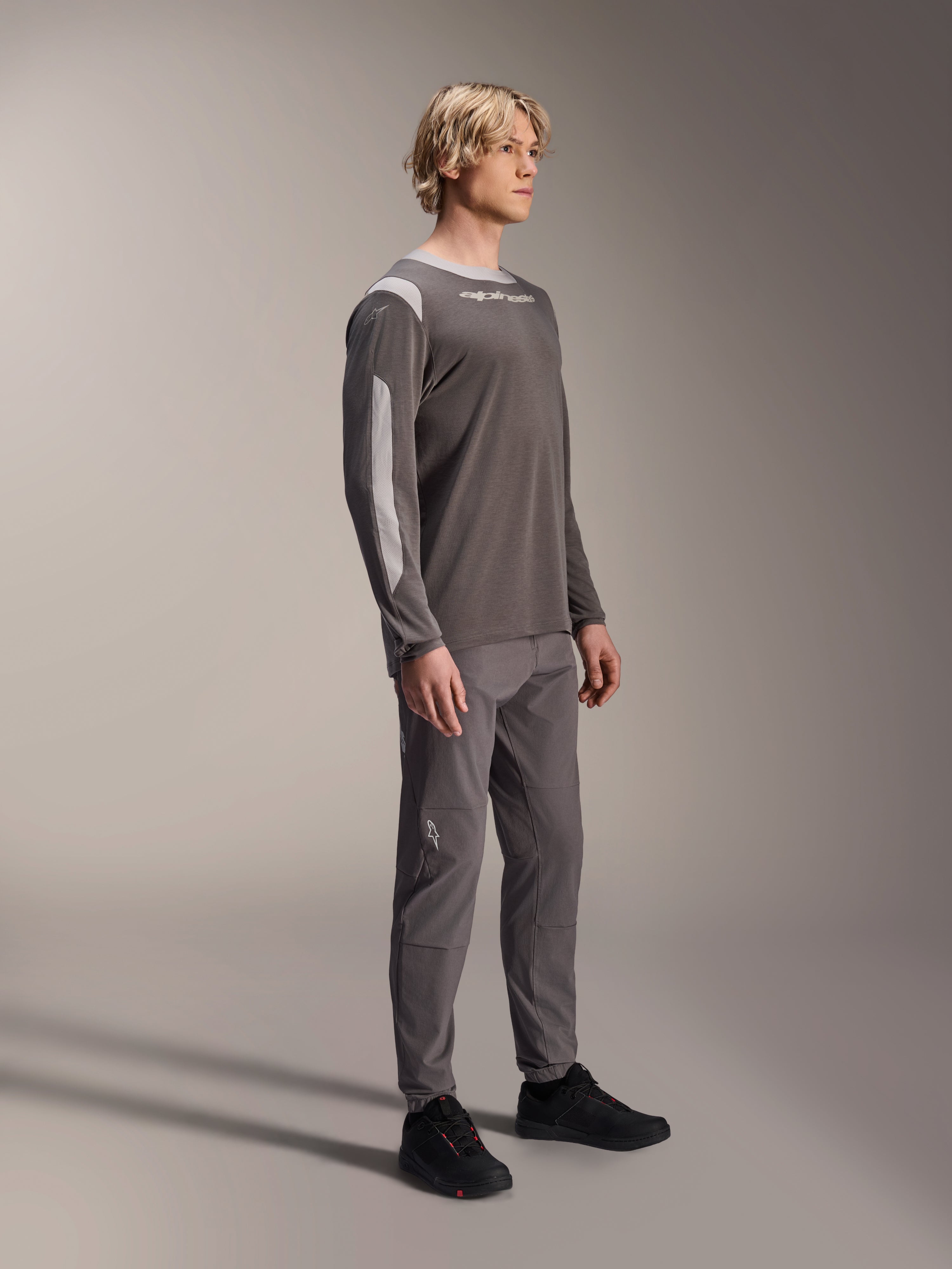 Męski model w koszulce Alpinestars A-Dura Dri Wool z długim rękawem w kolorze ciemnoszarym, zestawionej z pasującymi szarymi spodniami i czarnymi butami, jasnoszare panele z siateczki na ramionach i rękawach, techniczna odzież MTB do jazdy terenowej