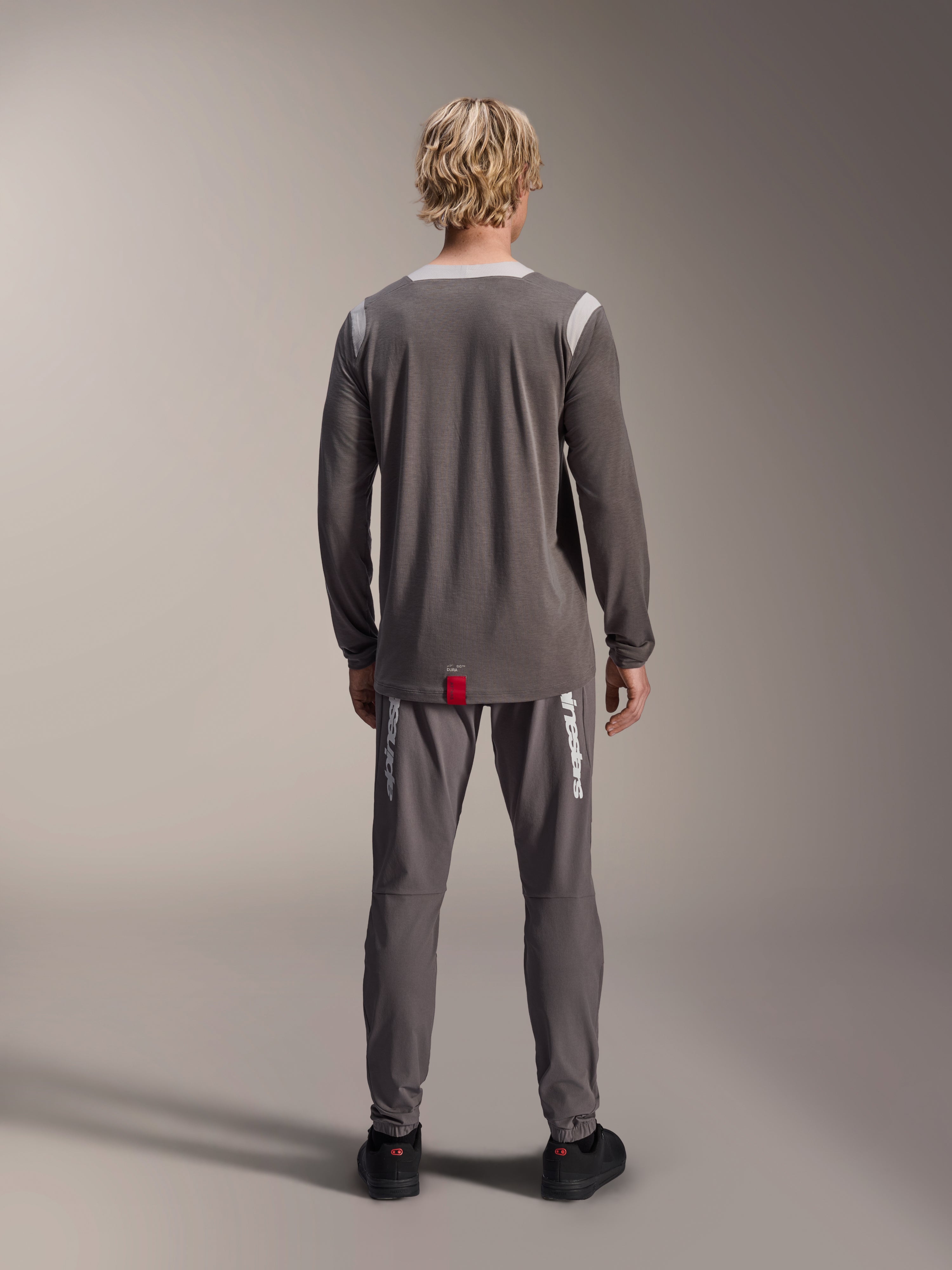 Koszulka Alpinestars A-Dura Dri Wool - długi rękaw, ciemnoszara, widok od tyłu na modelu, jasnoszare panele z siatki na ramionach i kołnierzu, przedłużony tył z małą czerwoną metką marki, zestawiona z pasującymi szarymi spodniami rowerowymi.