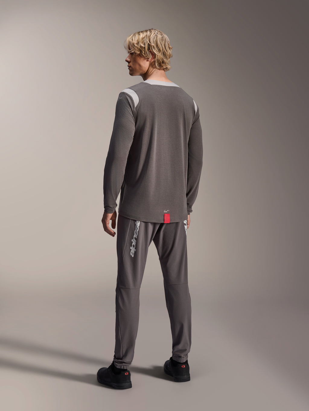 Męski model w koszulce Alpinestars A-Dura Dri Wool Long Sleeve, ciemnoszara z jasnoszarymi siateczkowymi panelami na ramionach, dopasowane ciemnoszare spodnie rowerowe, widok z tyłu prezentujący przedłużony tył, odzież MTB