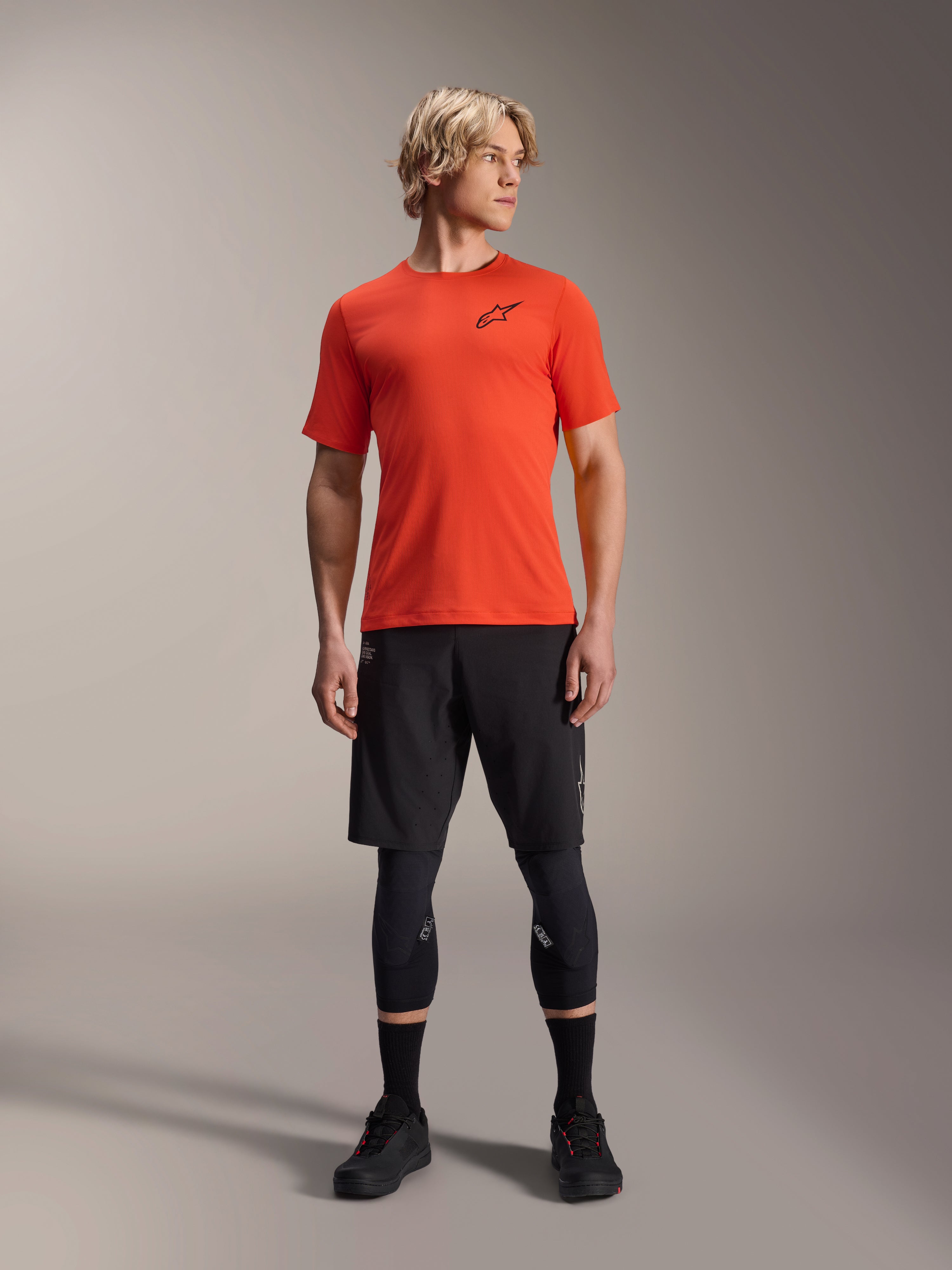 Koszulka z krótkim rękawem Alpinestars A-Aria Astar, Spicy Orange, noszona przez modela z czarnymi spodenkami MTB i ochraniaczami kolan, lekka i oddychająca odzież rowerowa do MTB