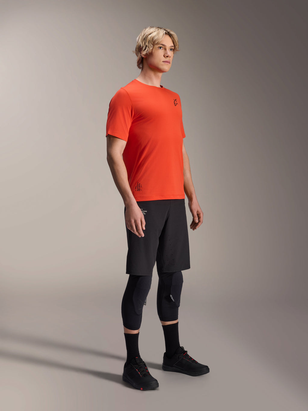 Koszulka Alpinestars A-Aria Astar z krótkim rękawem w kolorze Spicy Orange, prezentowana przez modela z czarnymi spodenkami MTB i ochraniaczami kolan, lekka oddychająca odzież na rower górski, intensywny pomarańczowy kolor z subtelnym czarnym logo
