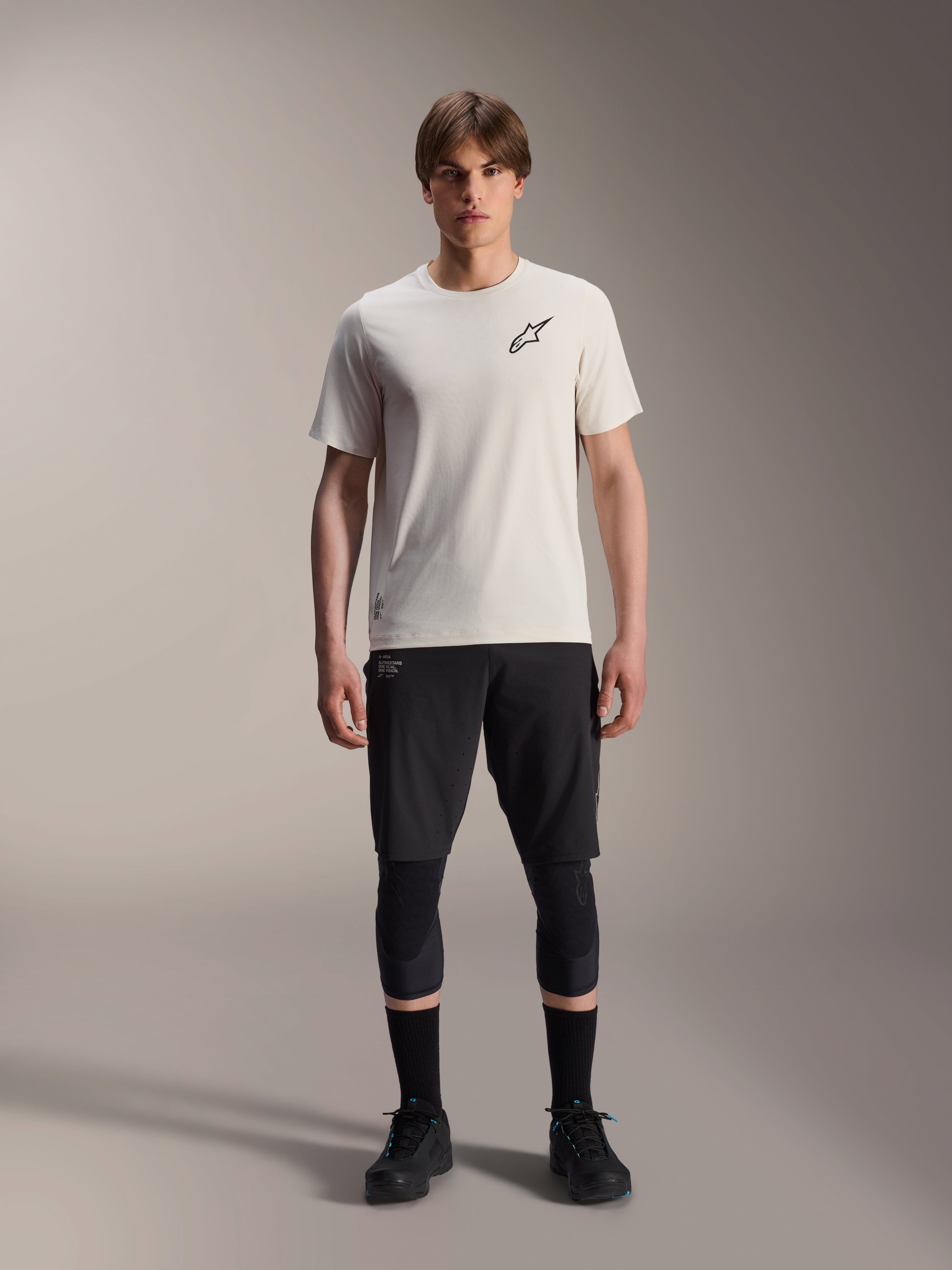 Męski model ubrany w koszulkę Alpinestars A-Aria Astar z krótkim rękawem w kolorze ecru, zestawioną z czarnymi szortami MTB, ochraniaczami kolan i czarnymi butami rowerowymi, z minimalistycznym czarnym logo gwiazdy na piersi, zaprojektowaną do jazdy terenowej na szlakach.