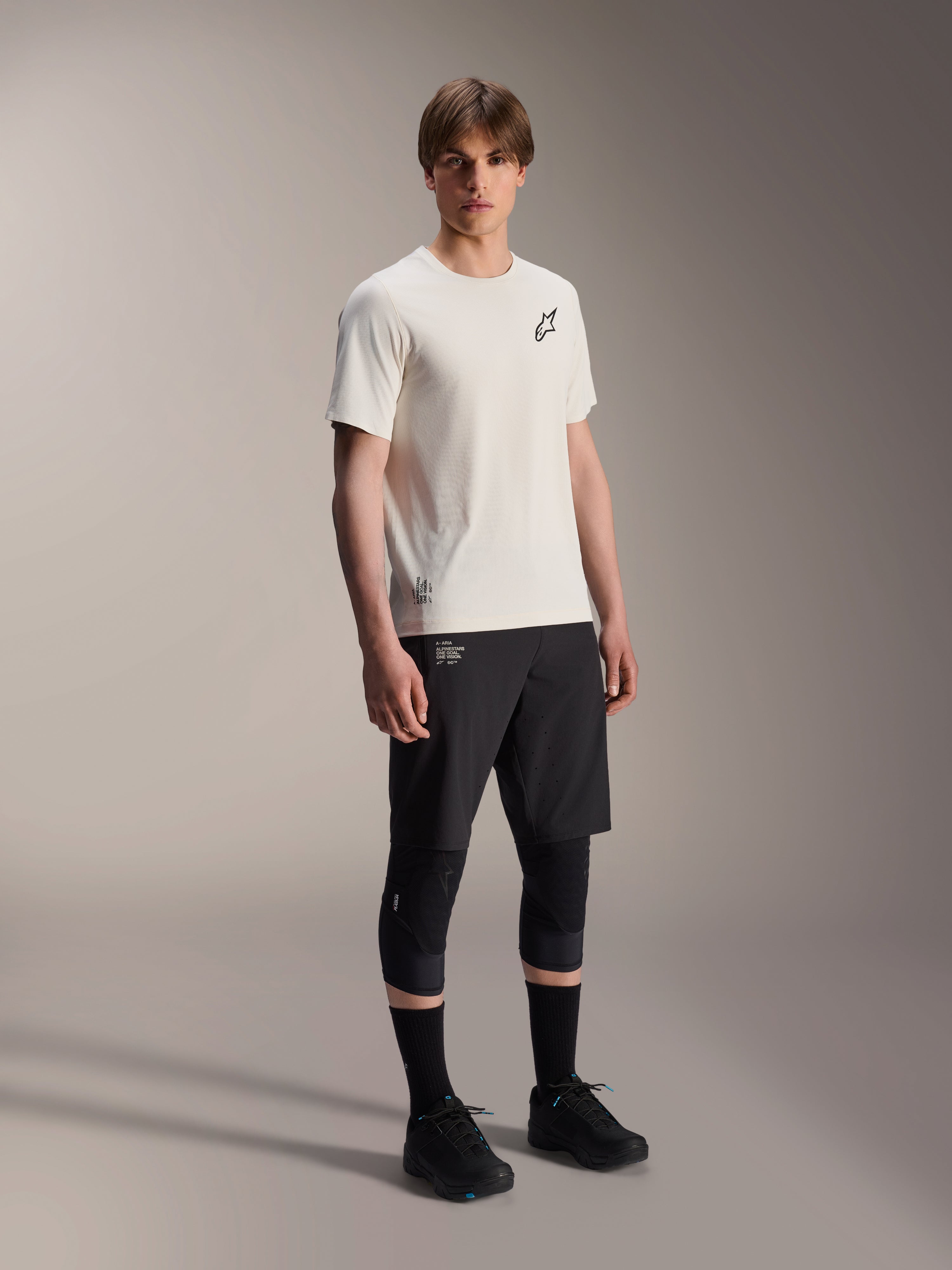 Alpinestars A-Aria Astar Jersey, koszulka z krótkim rękawem, kolor ecru, czarne logo na piersi, noszona z czarnymi szortami rowerowymi i ochraniaczami na kolana, lekka odzież wyczynowa MTB