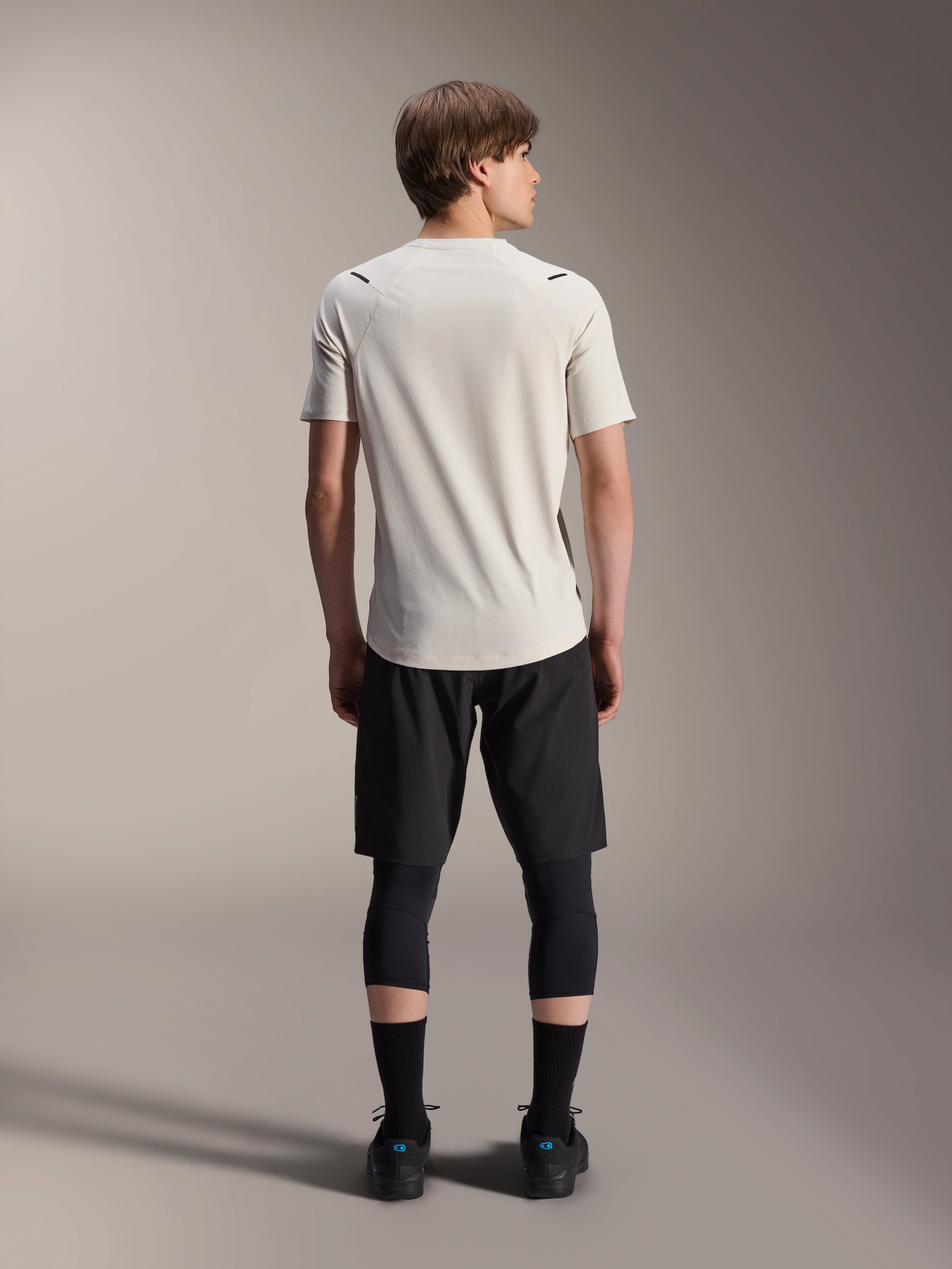 Męski model widoczny od tyłu w koszulce Alpinestars A-Aria Astar z krótkim rękawem w kolorze Ecru, czarne szorty MTB i legginsy ochronne, przedłużony tył i sportowy krój do jazdy w terenie.