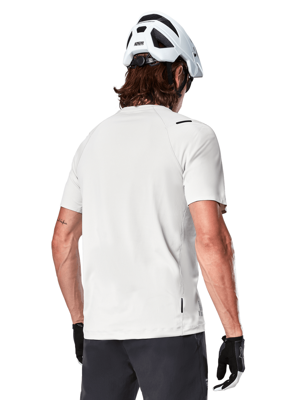 Alpinestars A-Aria Astar Jersey, krótki rękaw, kolor White Dove, widok z tyłu lekkiej i oddychającej koszulki rowerowej MTB, noszonej z białym kaskiem i czarnymi rękawiczkami rowerowymi, z wydłużonym tyłem dla lepszej osłony