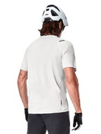 Alpinestars A-Aria Astar Jersey, krótki rękaw, kolor White Dove, widok z tyłu lekkiej i oddychającej koszulki rowerowej MTB, noszonej z białym kaskiem i czarnymi rękawiczkami rowerowymi, z wydłużonym tyłem dla lepszej osłony